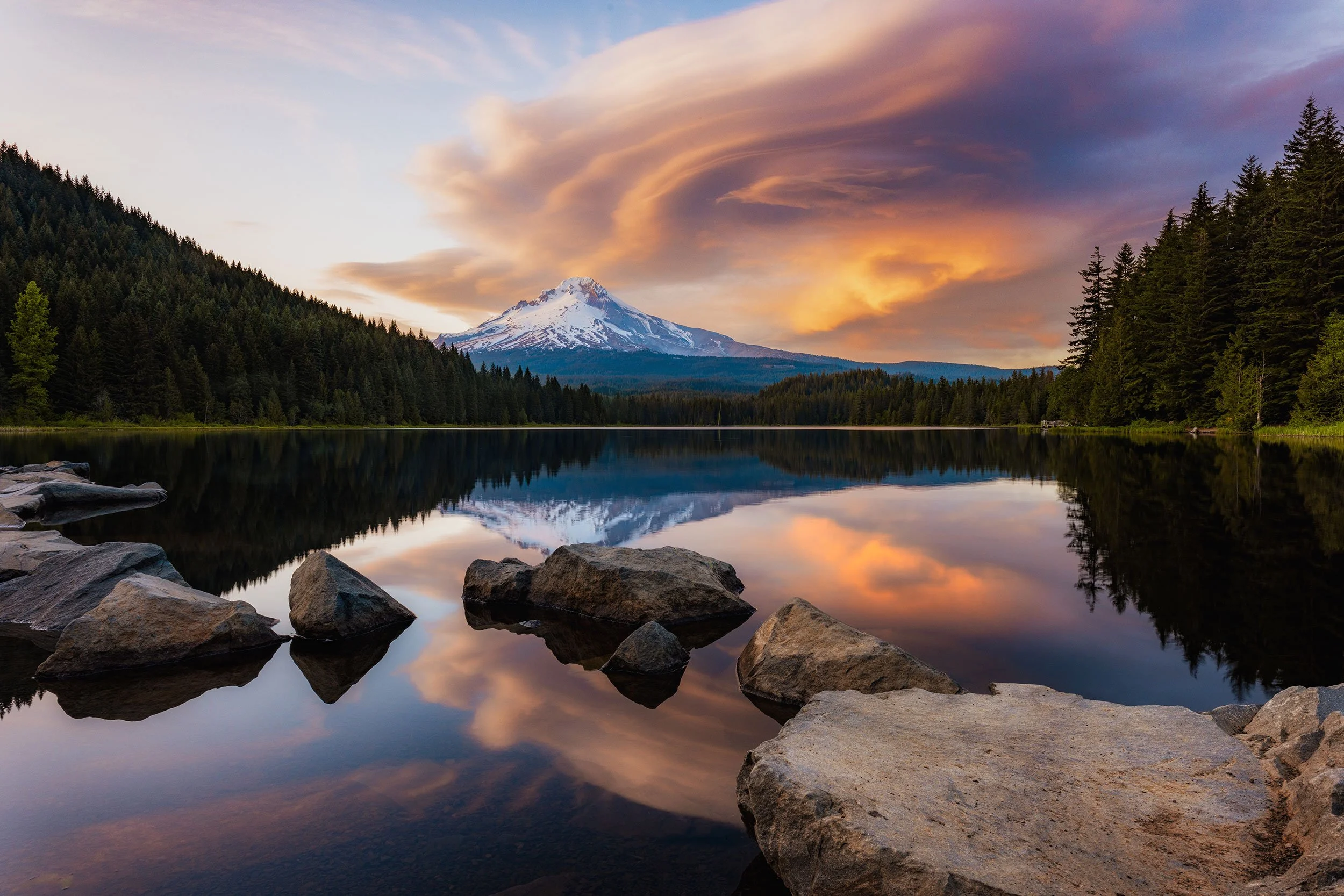 Fiery sunset and swirling lenticular clouds glowing above snow-capped Mount Hood, mirrored in the still water of Trillium Lake among foreground boulders, Oregon landscape photography by Ryan L. Smith
