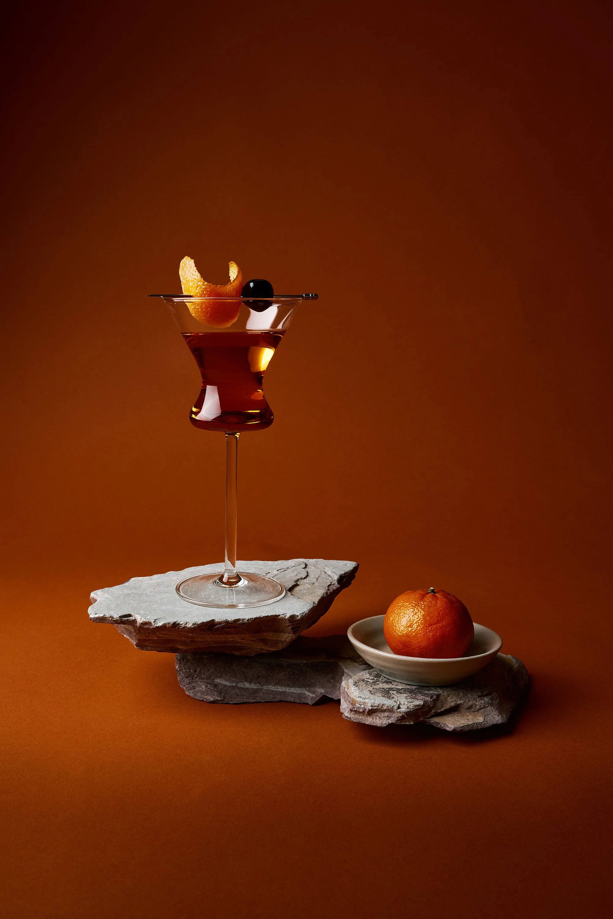 Styled Manhattan cocktail with orange peel and cherry garnish on stone slab, beverage photography by Ryan L. Smith