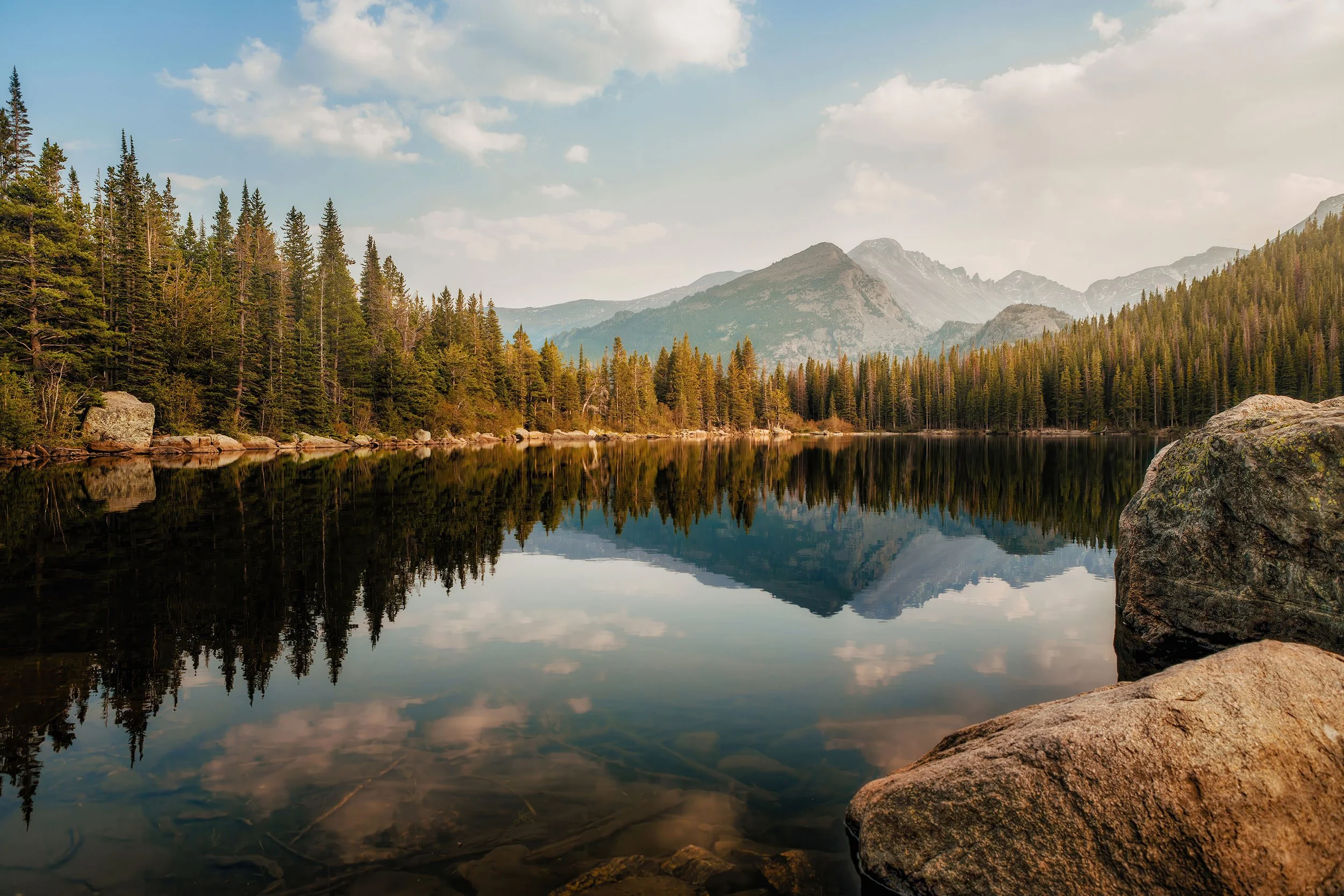 Landscape photograph of a calm alpine lake surrounded by pine trees and mountains in Rocky Mountain National Park.