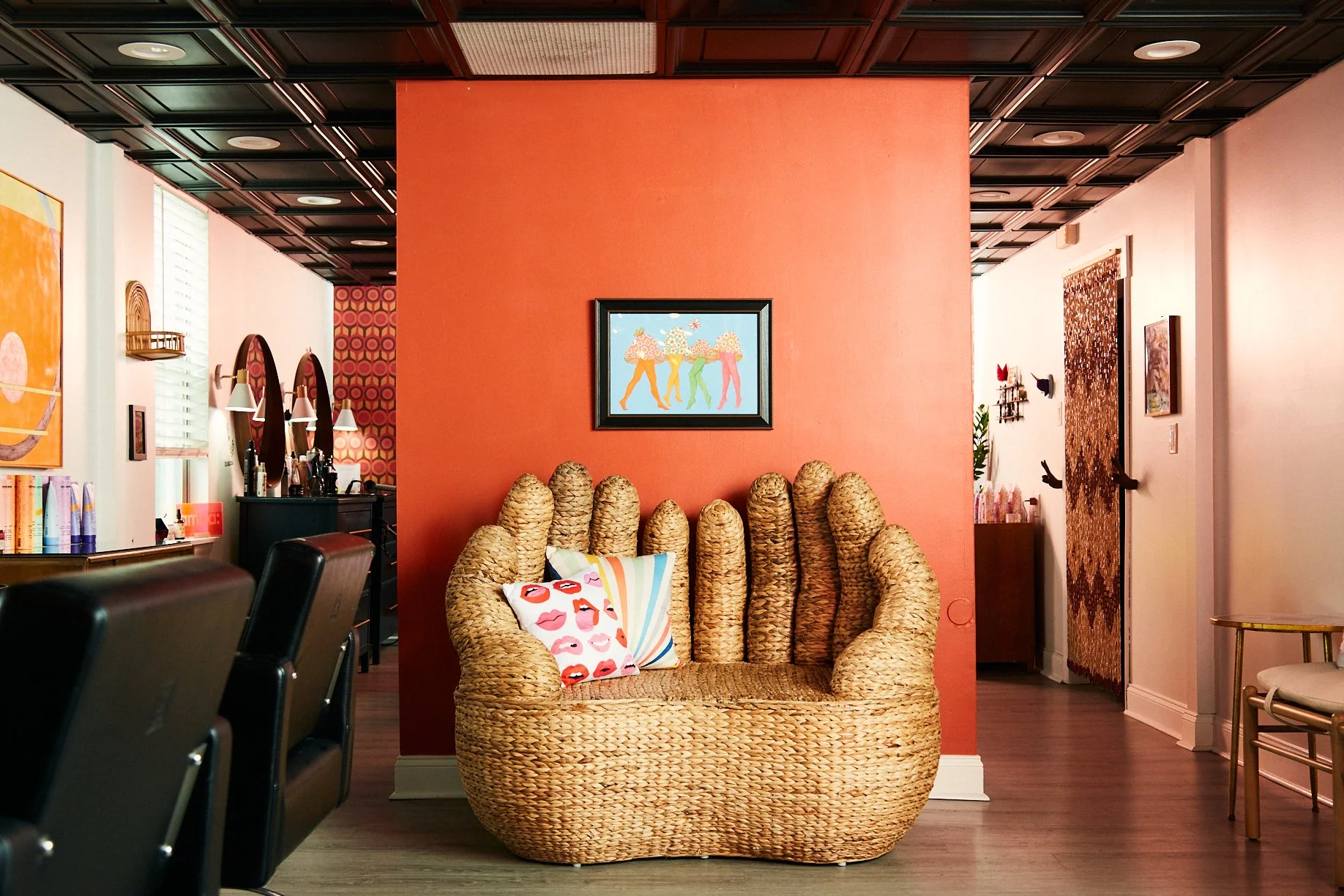 Interior of a colorful, cozy room with a woven wicker sofa that has two decorative pillows, one with lips print and the other with rainbow stripes, placed against a coral wall. To the left, there are chairs and a mirror with a makeup station. On the right, a doorway with a patterned curtain is visible, along with wall decorations and small furniture pieces.