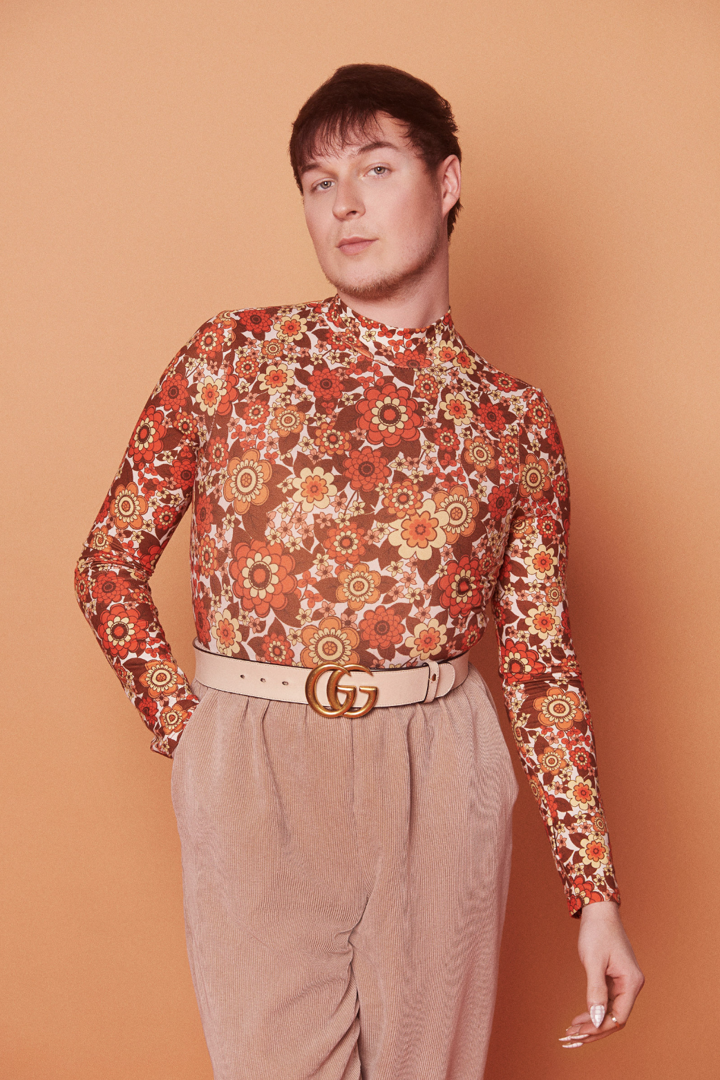 A person wearing a long-sleeve floral shirt with red, orange, and yellow flowers, beige high-waisted pants, and a white belt with a gold Gucci logo buckle, standing against a peach-colored background.