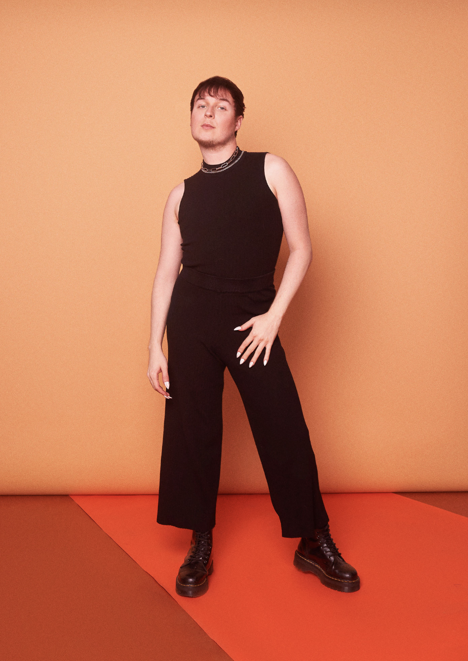 Person standing in front of an orange background, wearing a black sleeveless top, black wide-leg pants, and black boots, with hand on hip and a confident pose.