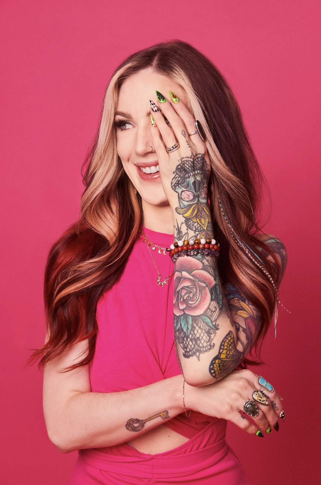 A young woman with long, wavy, two-toned hair (blonde and dark brown) stands against a pink background. She is smiling and playfully covering her right eye with her right hand. She is wearing a bright pink top, several rings, and multiple bracelets. She has colorful tattoos on her right arm and hand, including roses, a butterfly, and a key. Her nails are decorated with bright, patterned nail art.