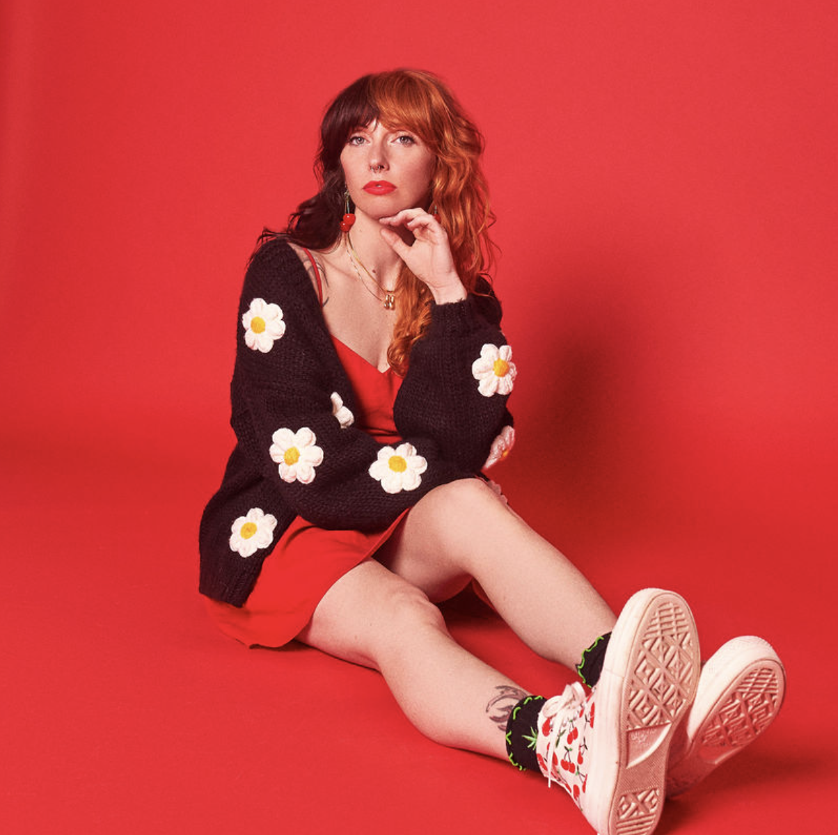 A woman with red hair, sitting against a red background, wearing a red dress, a black sweater with white daisies, white sneakers with red cherries, and floral socks, with her chin resting on her hand.