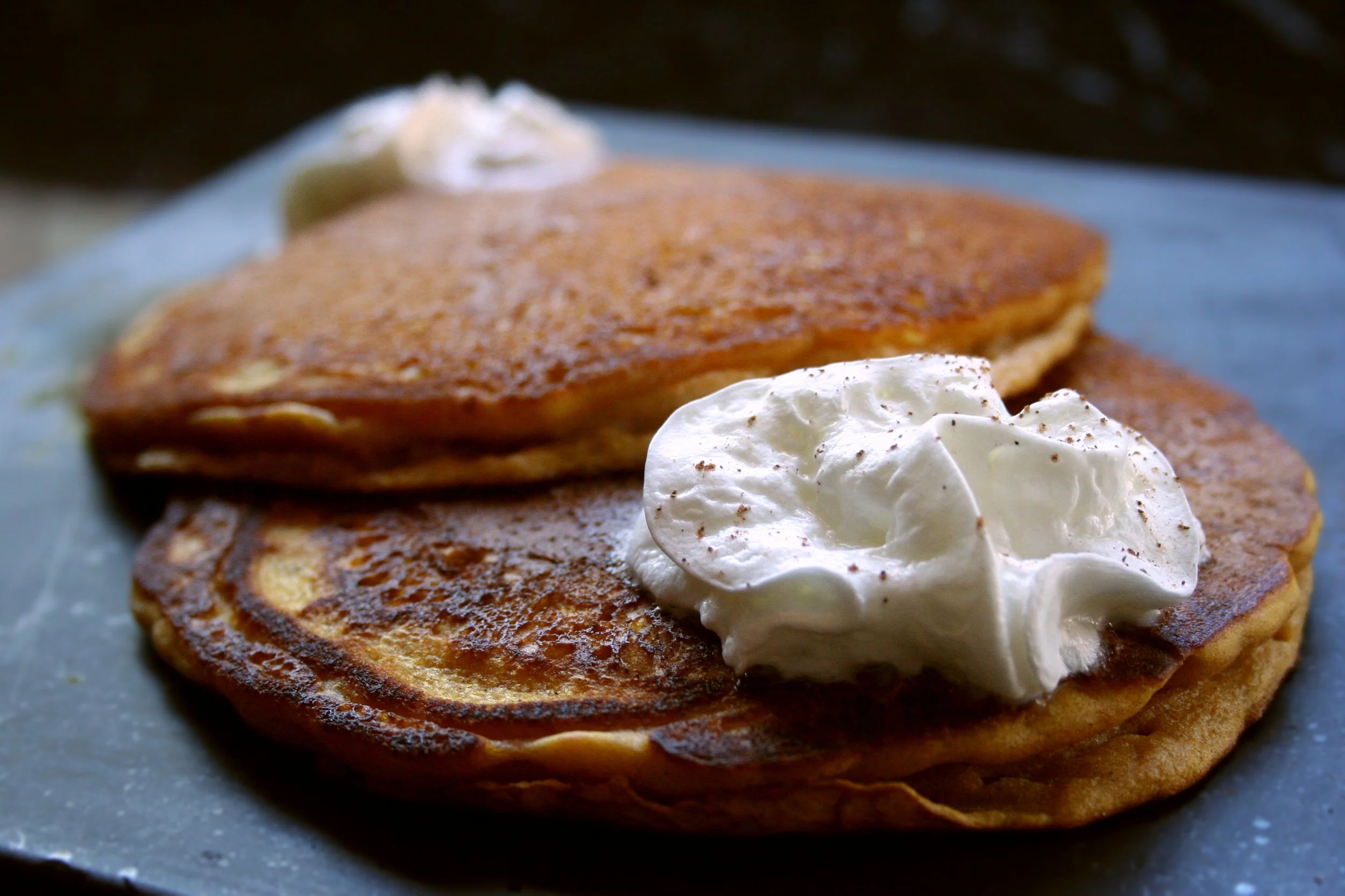 Close-up of two stacked golden-brown pancakes topped with whipped cream and a dollop of butter on a dark plate.