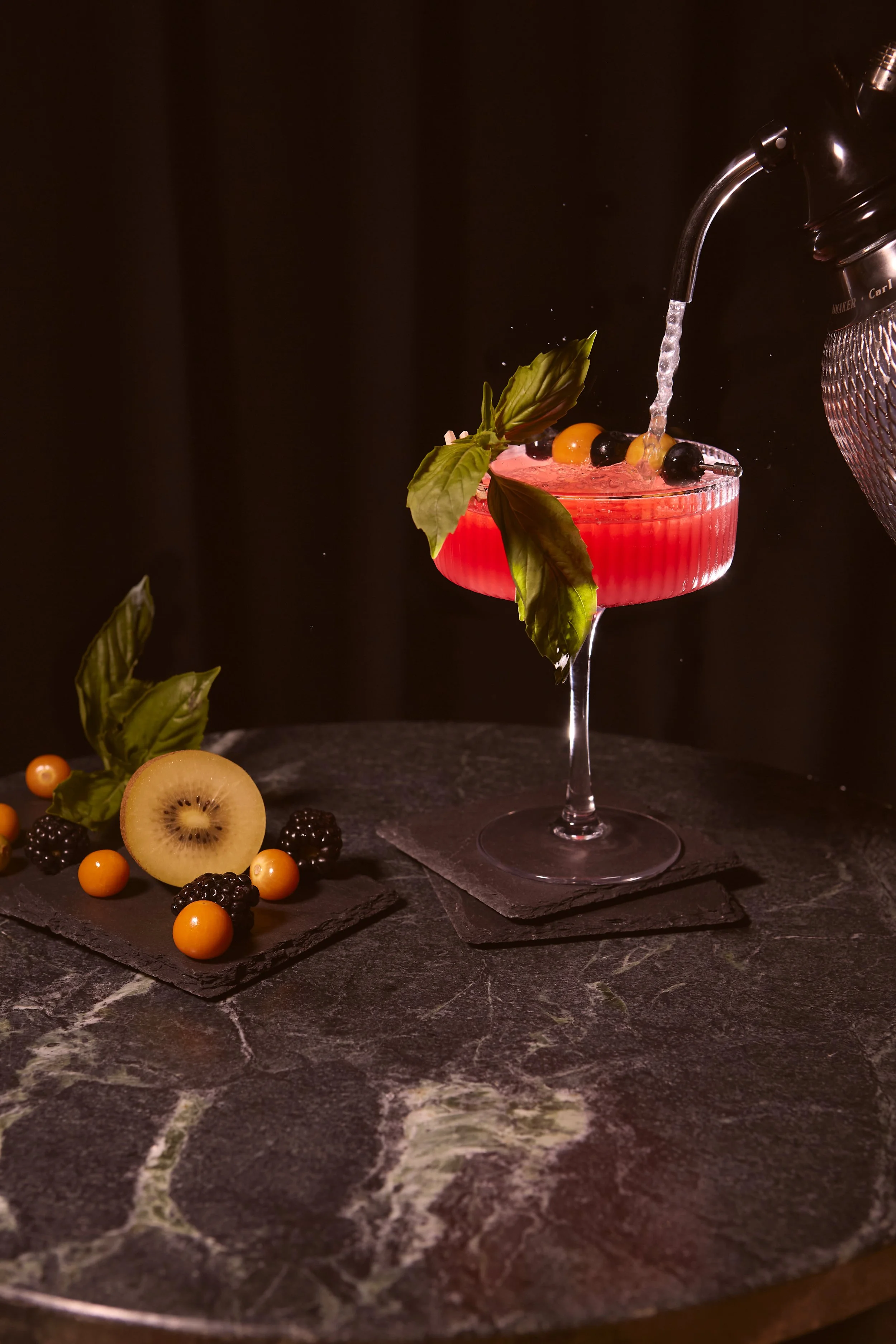 A pink cocktail garnished with basil leaves and blackberries on a dark background, with scattered berries and a slice of kiwi on a marble surface.