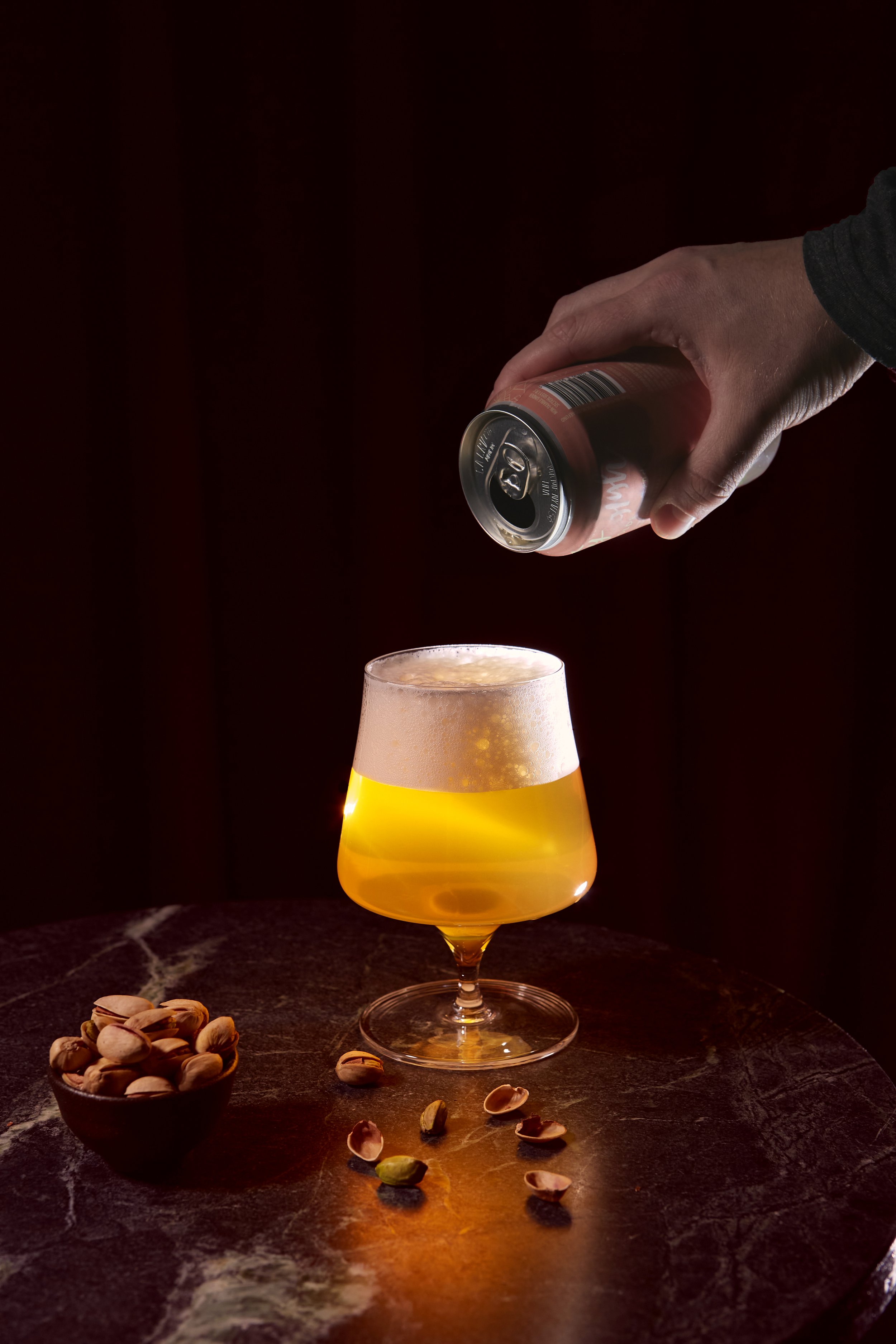 A hand pouring a canned beverage into a tulip-shaped glass with a foamy head, a bowl of pistachios on the dark marble table.