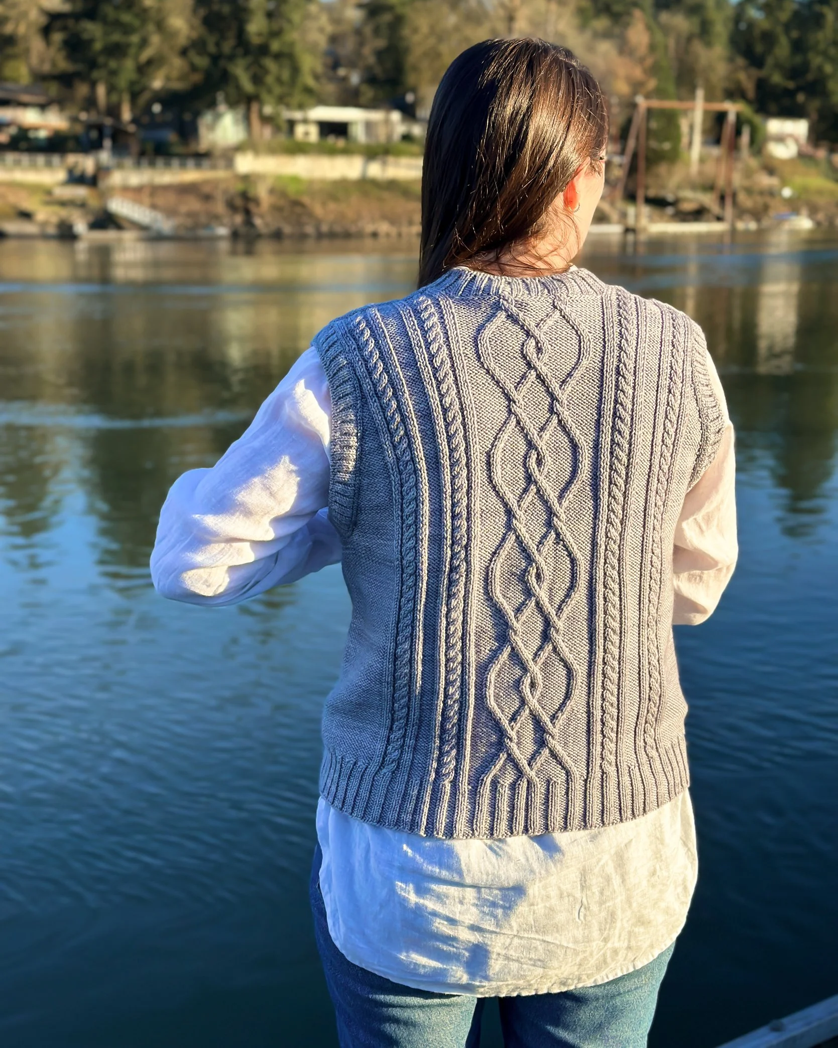 Cables really stand out on the back of Tryon Creek Vest as a beautiful young woman wears it beside the river as the sun goes down.