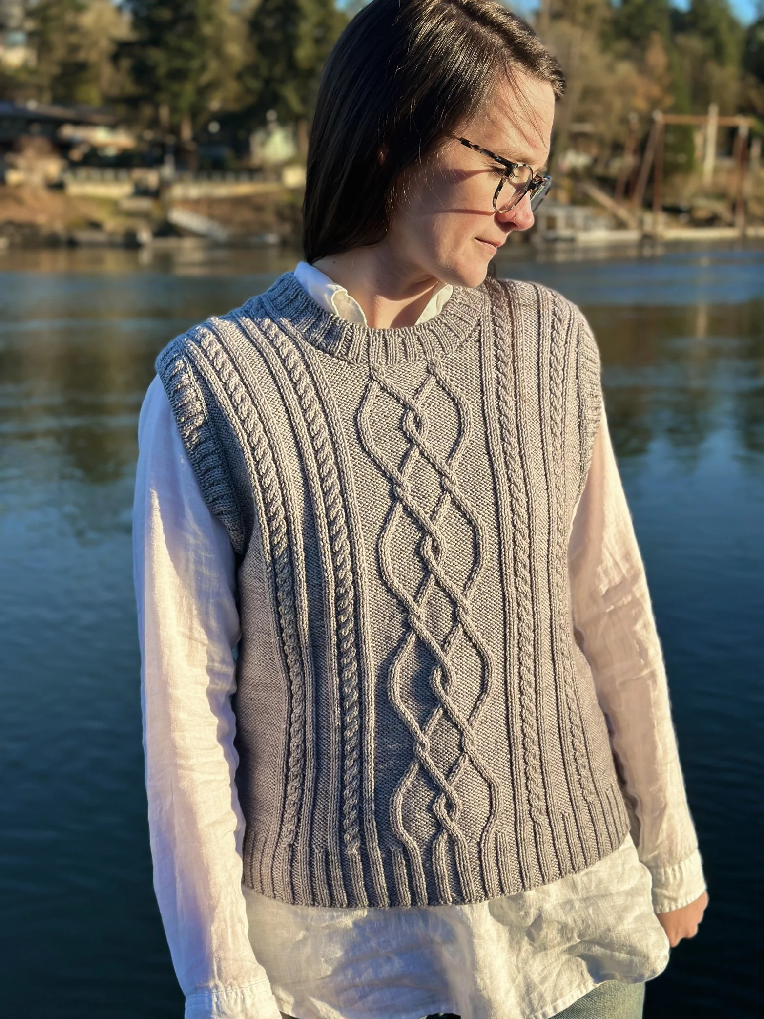Tryon Creek Vest - Model is wearing silvery grey cabled vest by the river. Sample is knit in @madelinetosh Tosh DK yarn.