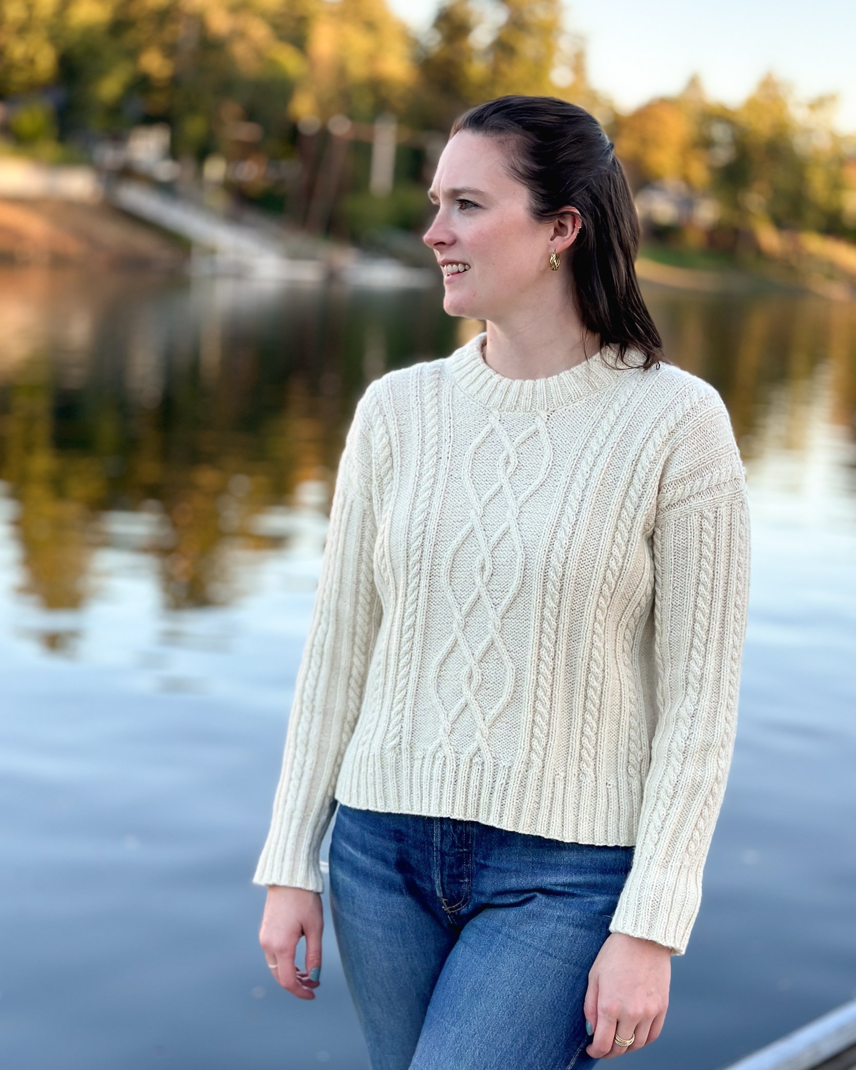 Tryon Creek Knit Sweater - Model is wearing sample by river in front of fall foliage. Yarn used is The Fibre Co Cumbria in color White Heather.