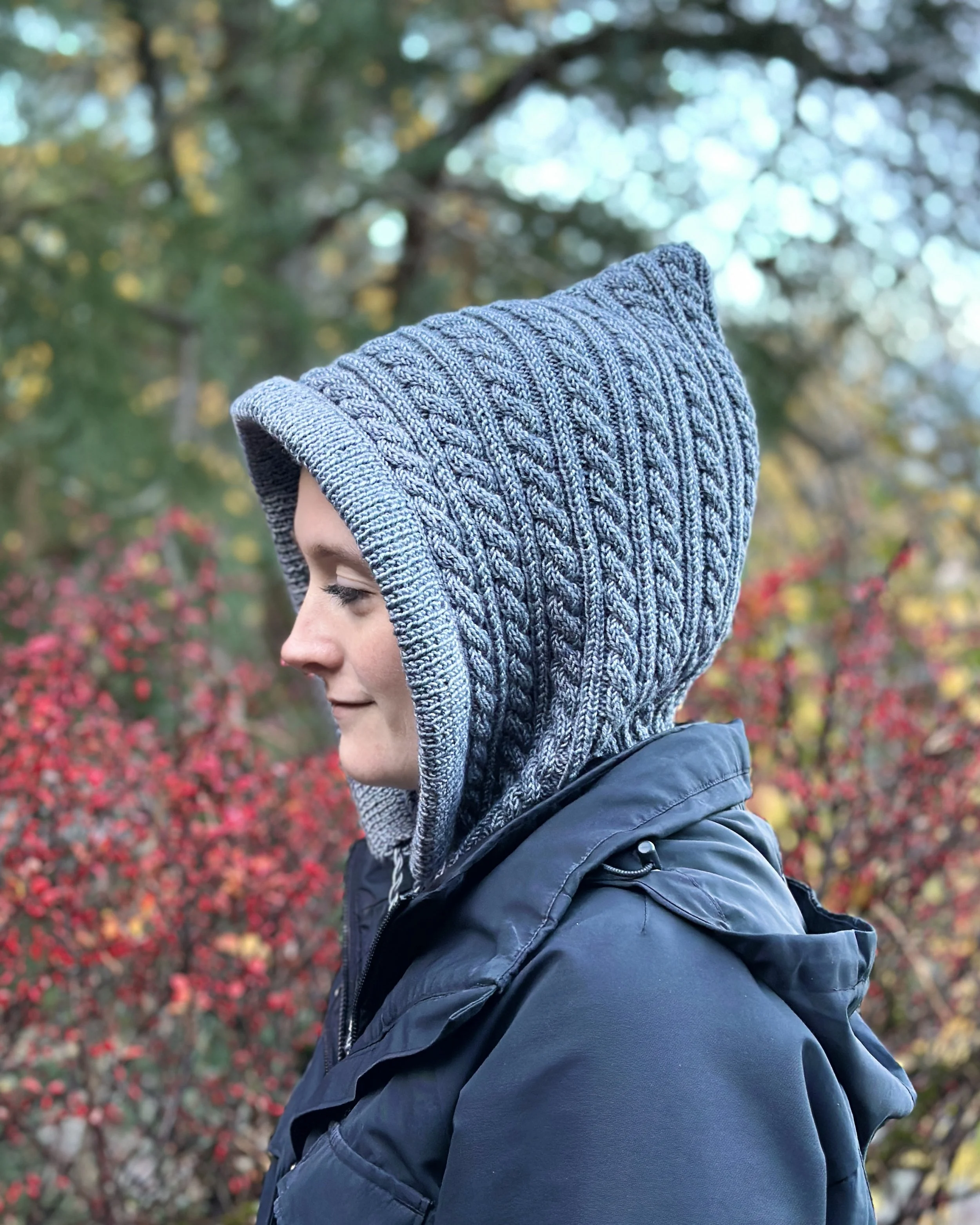 Tryon Creek, an easy cabled hoodie/cowl (balaclava) knitting pattern, that can be worn as a cowl. Model smiling while wearing a blue-grey version of Tryon Creek Hoodie with hood up.