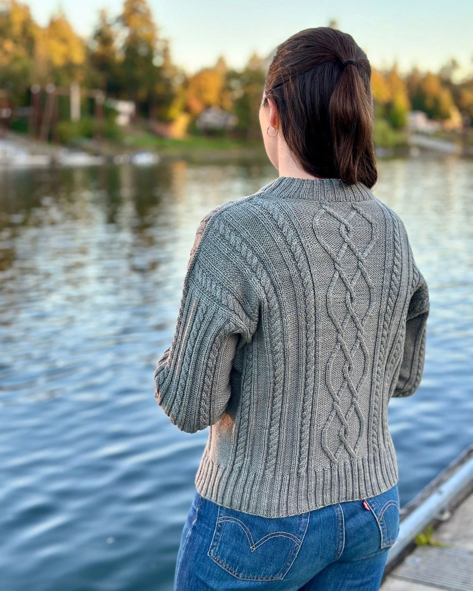 Tryon Creek Knit Sweater - Back of sweater on model by river with autumn foliage in background. Yarn used is Miss Babs Killington in color Arthurs Seat.
