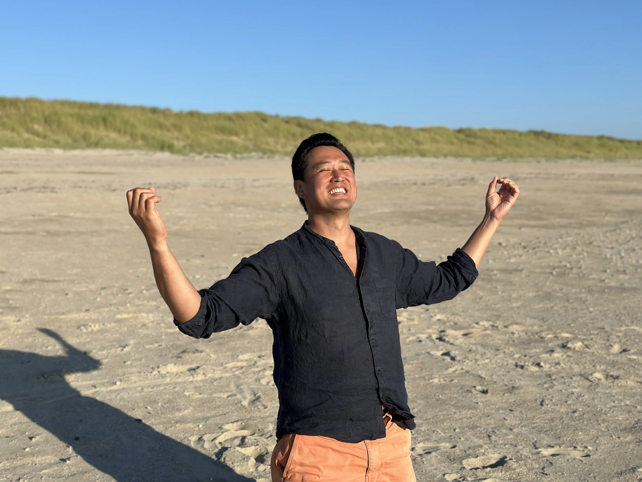 Joo Teoh, author, acupuncturist, yoga and qigong teacher is standing on a beach with his arms outstretched, an elated expression on his face.