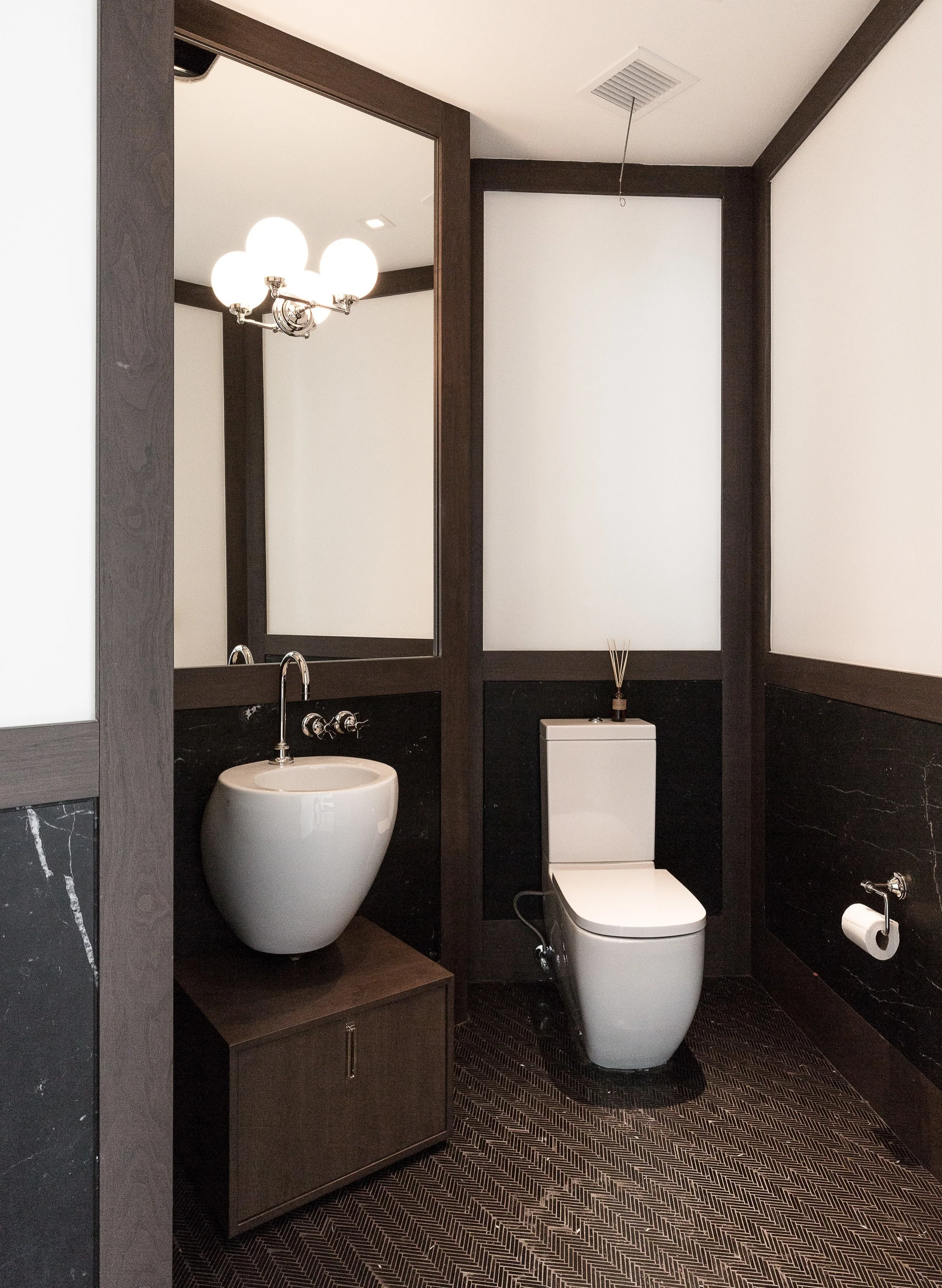 The chevron tiles in this powder room and natural stone panels give it a refined vintage look, which is complemented by the globe lighting fixtures. Design by ODA Architecture