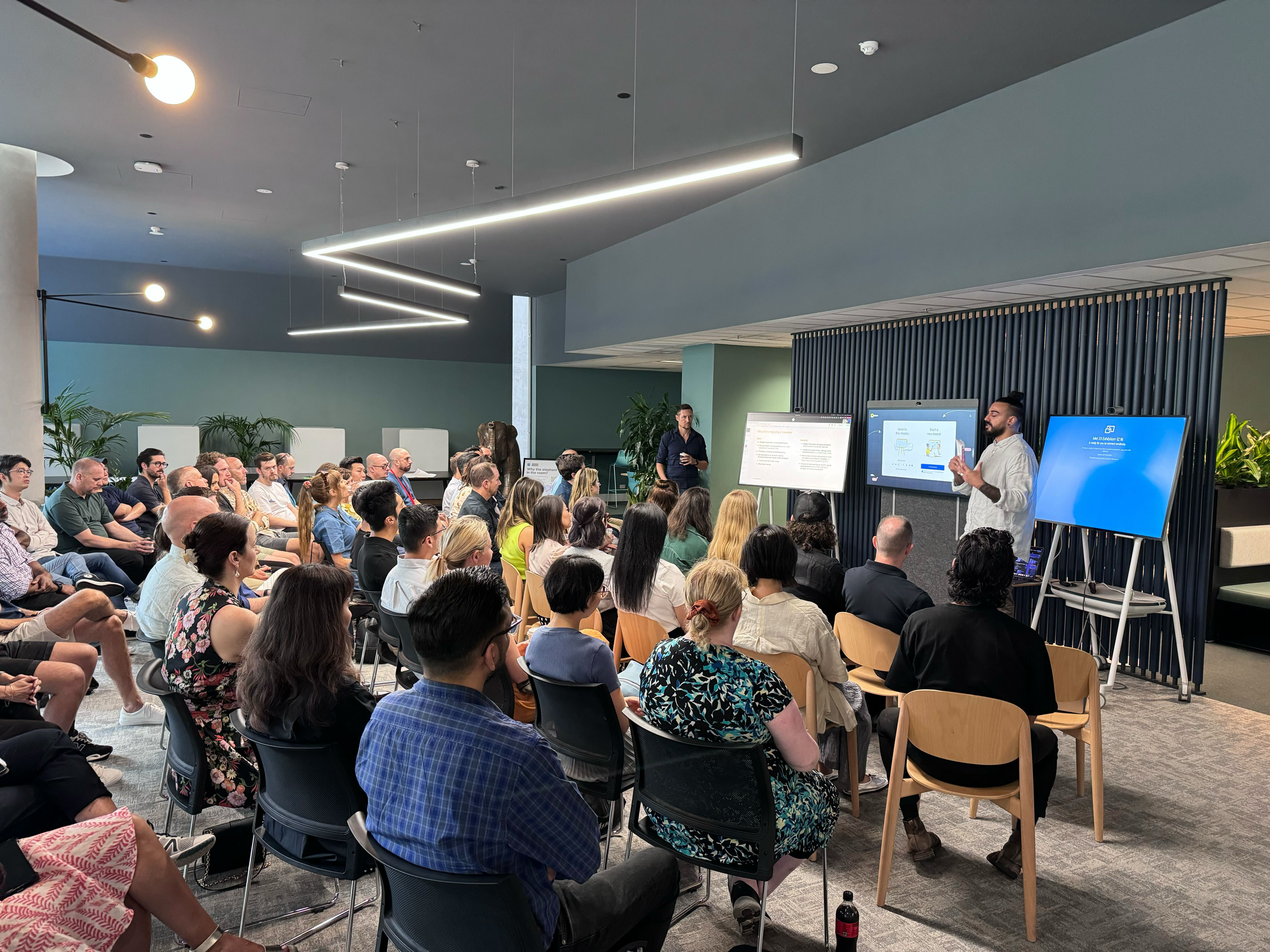 A facilitator presenting to a large, seated audience in a modern office space, leading a strategic facilitation and training session to unlock a full design function for a Tier 1 healthcare provider.