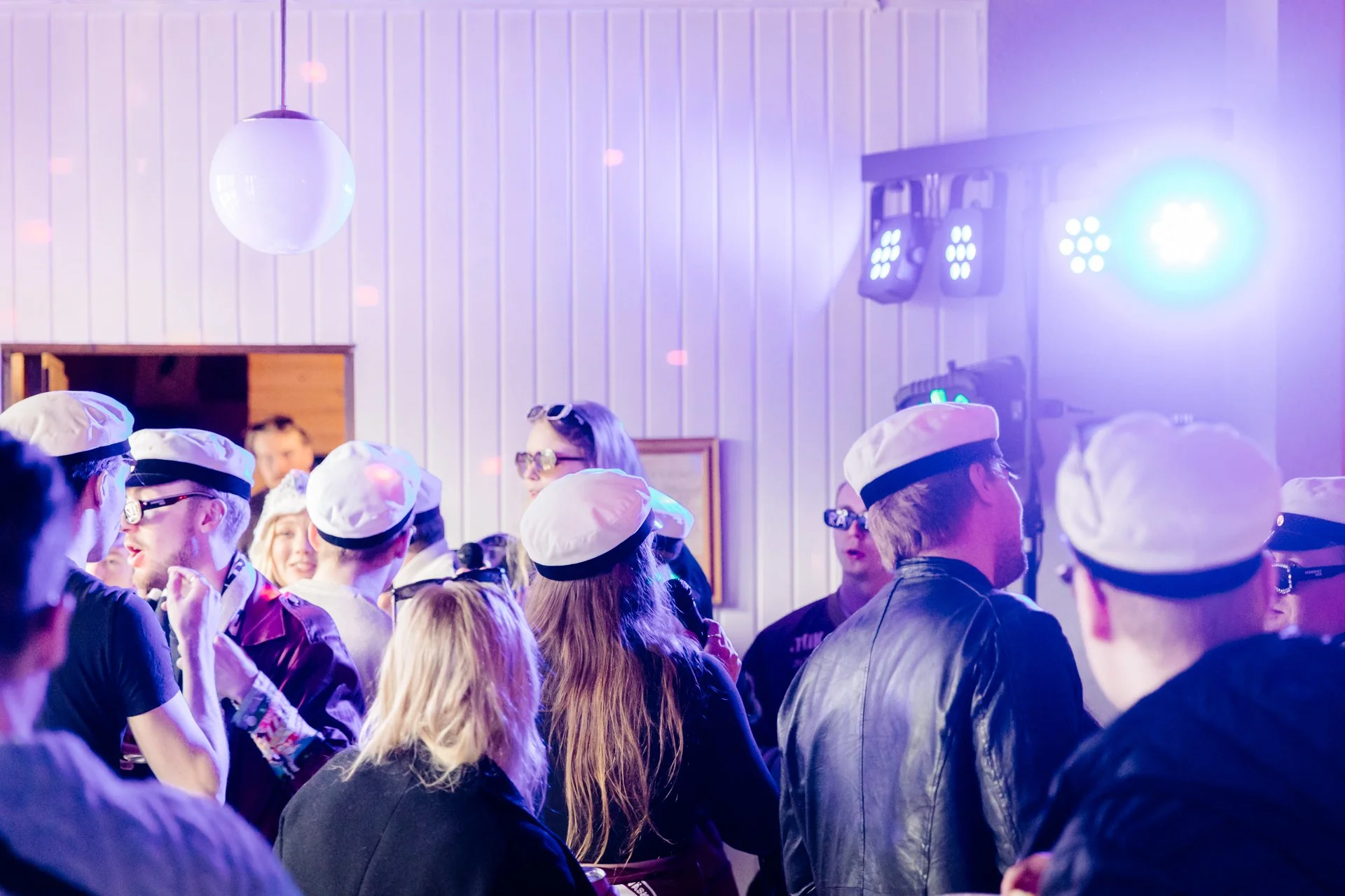 Nuori väki keskustelee illan vietosta, osa on pukeutunut juhlapukuihin ja päähineisiin, taustalla DJ:n soittamat musiikit ja valotehosteet.