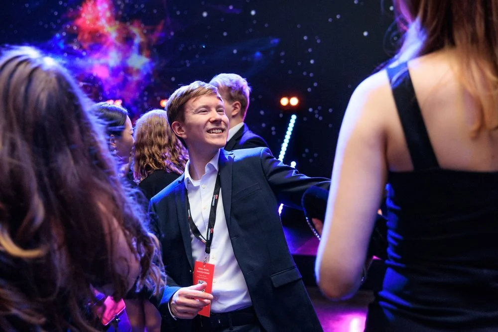 Nuori mies hymyilee ja tanssii juhlatilassa muiden ihmisten ympärillä, taustalla näkyvä galaxymaisesti valaistu tausta.