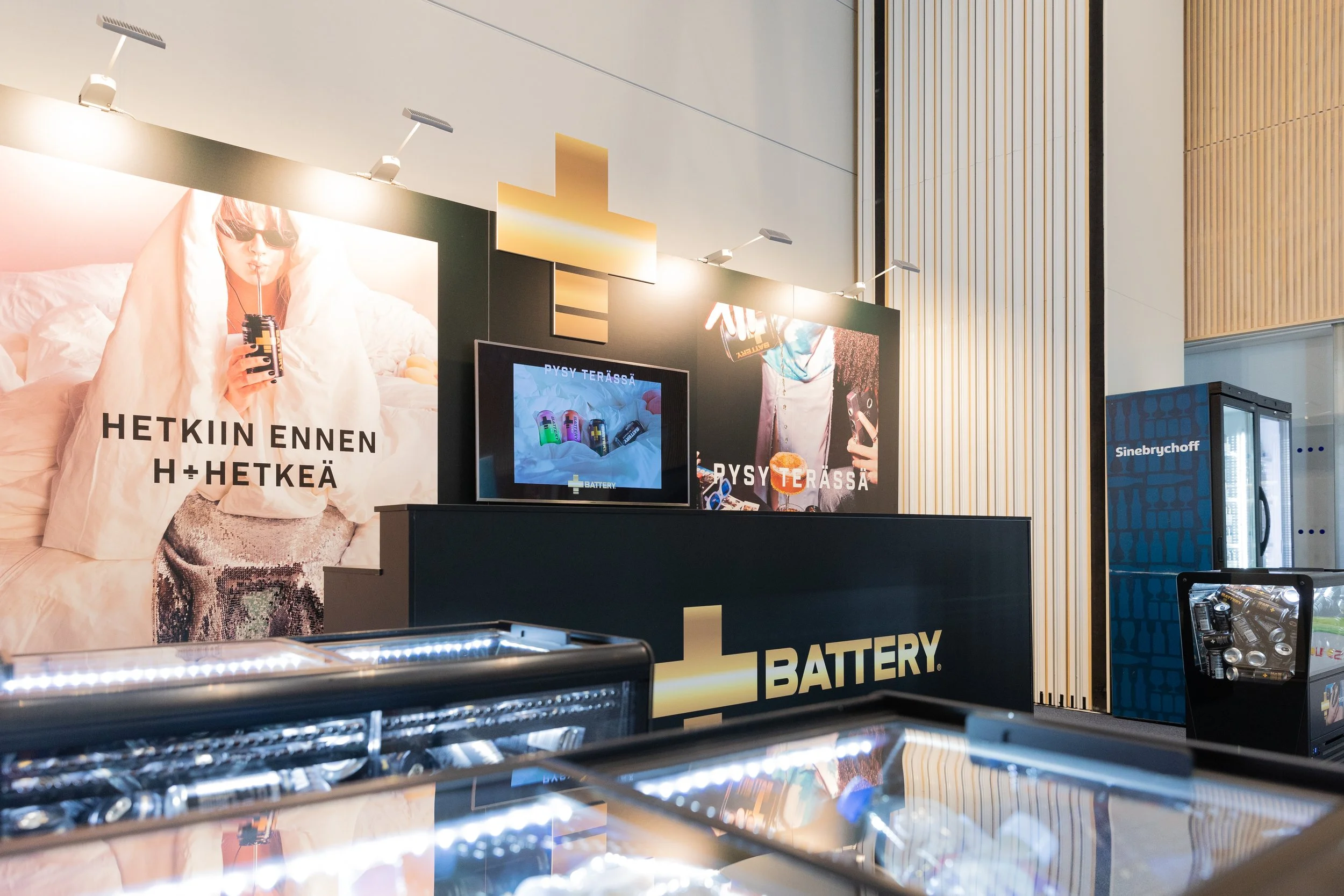 Näyttelytila battery- ja Sinebrychoff-tuotteille, esillä posterit ja jäähdyttimet, joissa on tuotteita, ja myyntitiski.