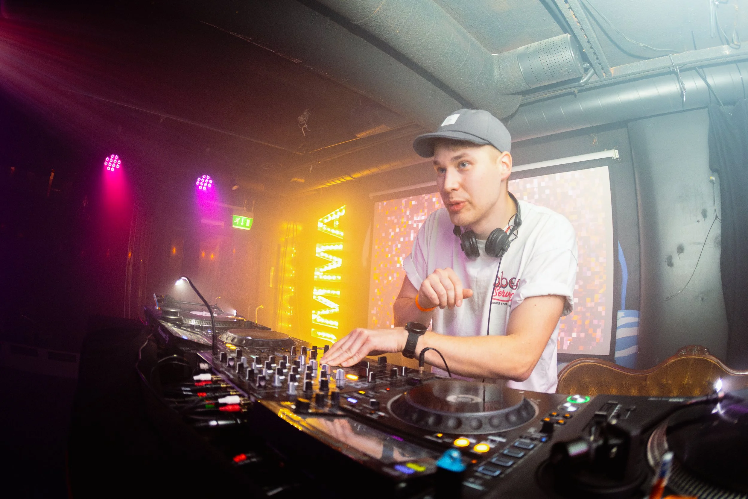 DJ soittaa musiikkia klubilla, taustalla värikkäitä valoja ja suuri esiintymissäteel oleva 'LIVE' -kyltin valot.