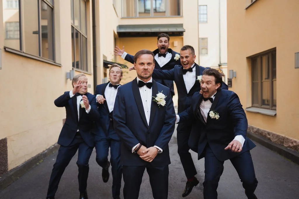 Seitsemän miestä pukeutuneina juhla-asuihin, osa tanssii ja naureskelee, taustalla vaaleansiniset rakennukset.