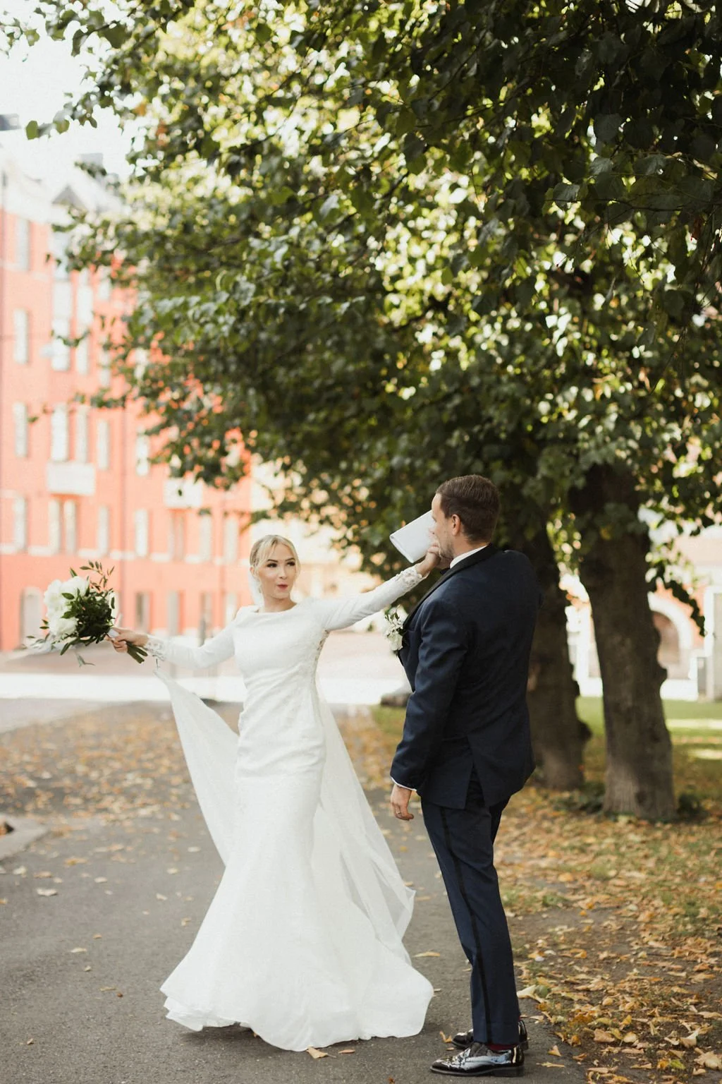 Hääparin välinen leikki, jossa nainen ja mies ovat ulkona puiden alla, nainen heittää kukkakimpun miehelle, joka vastaa sitä, ja nainen on pukeutunut valkoiseen hääpukuun, mies tummansiniseen pukuun. Taustalla puut ja rakennuksia.