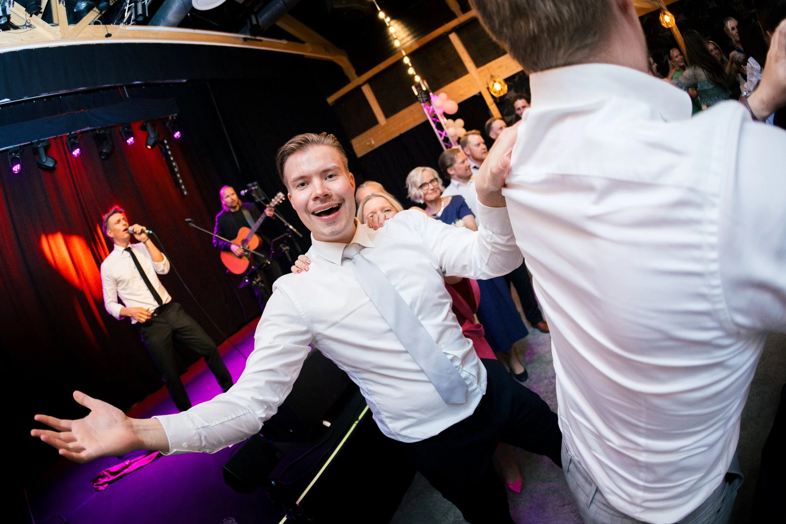 Hymyilevä nuori mies tanssimassa juhlassa, taustalla esiintyy laulaja ja kitaristi.