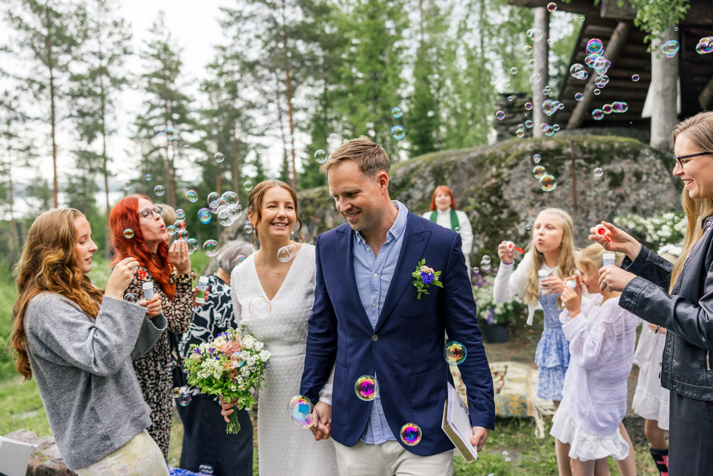 Hääparin käsi kävelee tai kulkee kohti kameran etualalla, taustalla onnellinen hääjuhla, ilmapalloja ja saippuakuplia, ulkona metsän ympäröimänä, hääparin ja vieraiden ympärillä.
