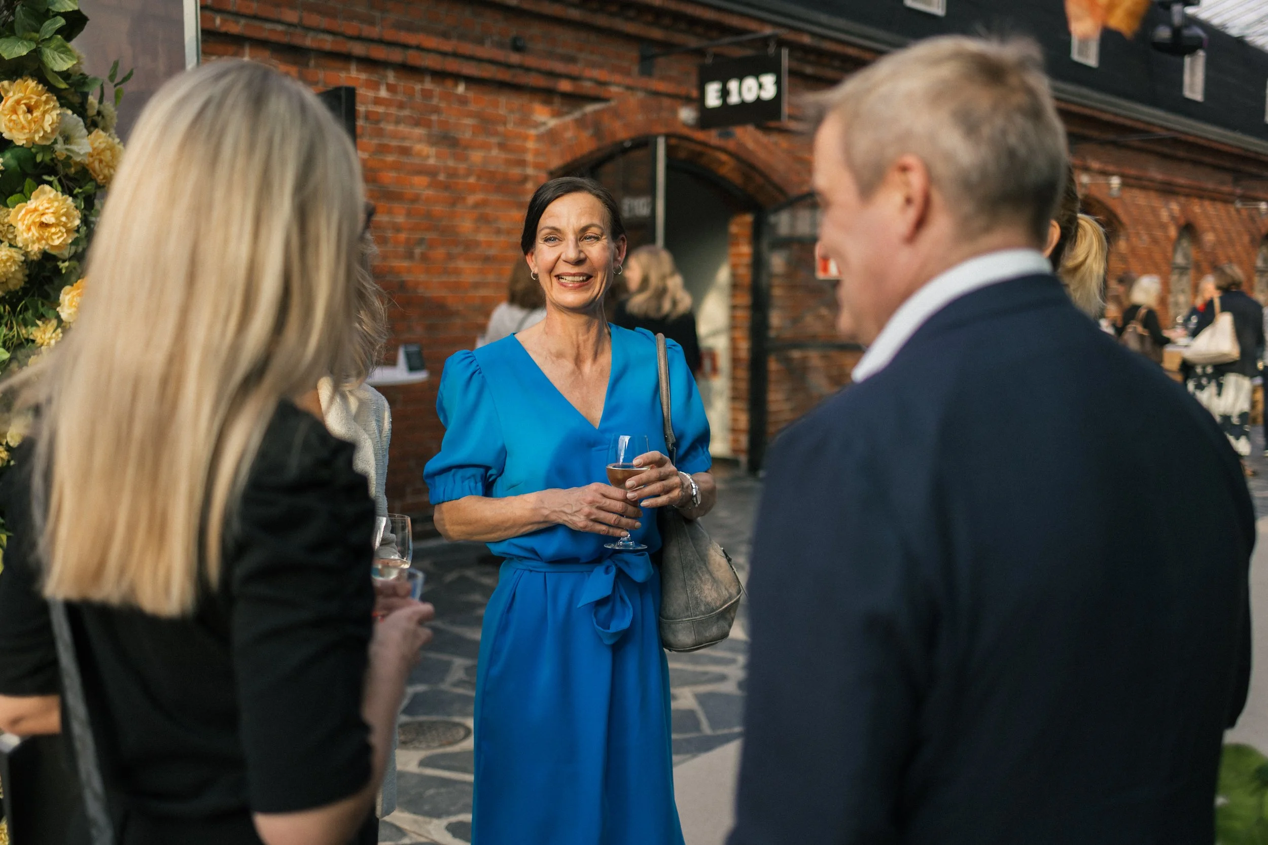 Nainen puvussa, sininen mekko, hymyilee ja pitää lasillista, keskustelee kahden muun ihmisen kanssa ulkona, taustalla punainen tiiliseinä.