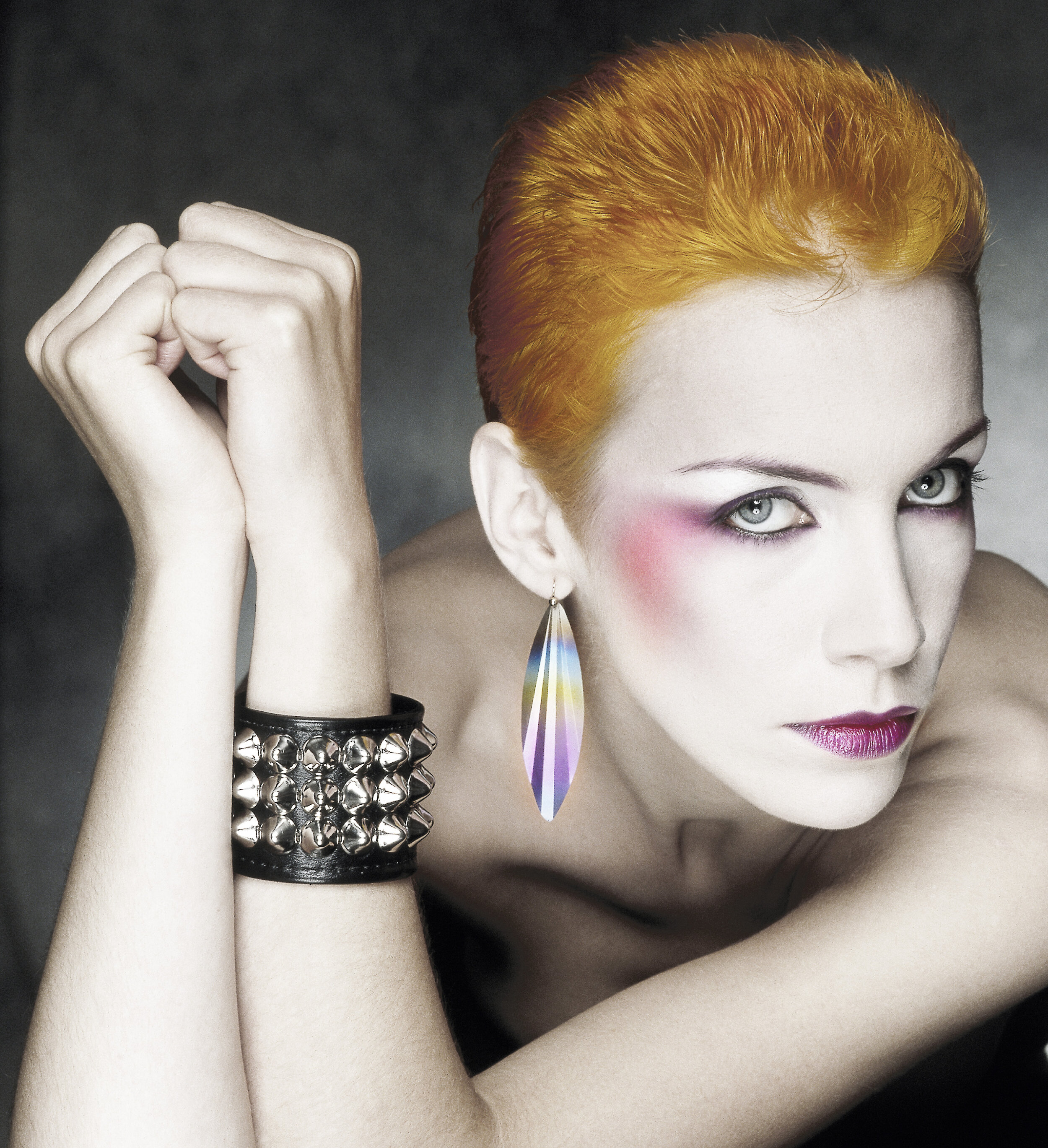 Annie Lennox with orange hair and pale white face and make up looking at camera. Her arms are rested on a box and she is leaning behind them