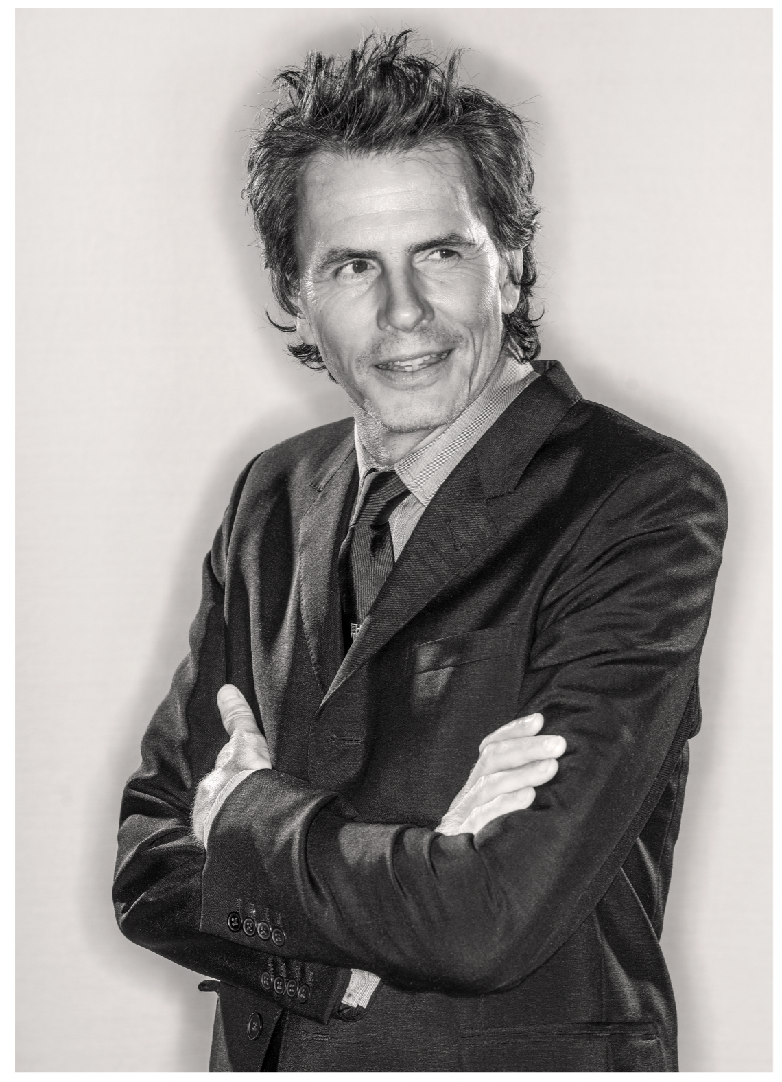 John Taylor of Duran Duran wearing suit and tie with arms folded looking off camera and smiling.