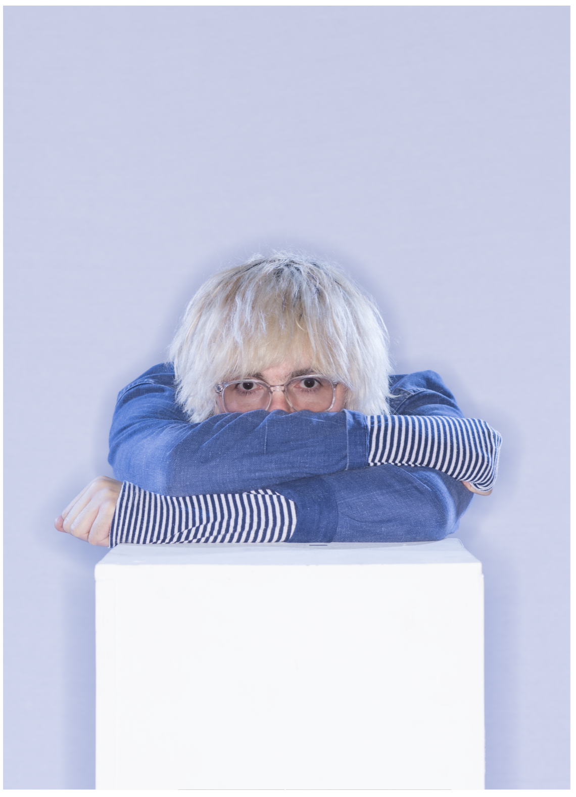Tim Burgess, The Charlatans wearing a blue top with a stripey t shirt leans over white box his face partially hidden by his arms.
