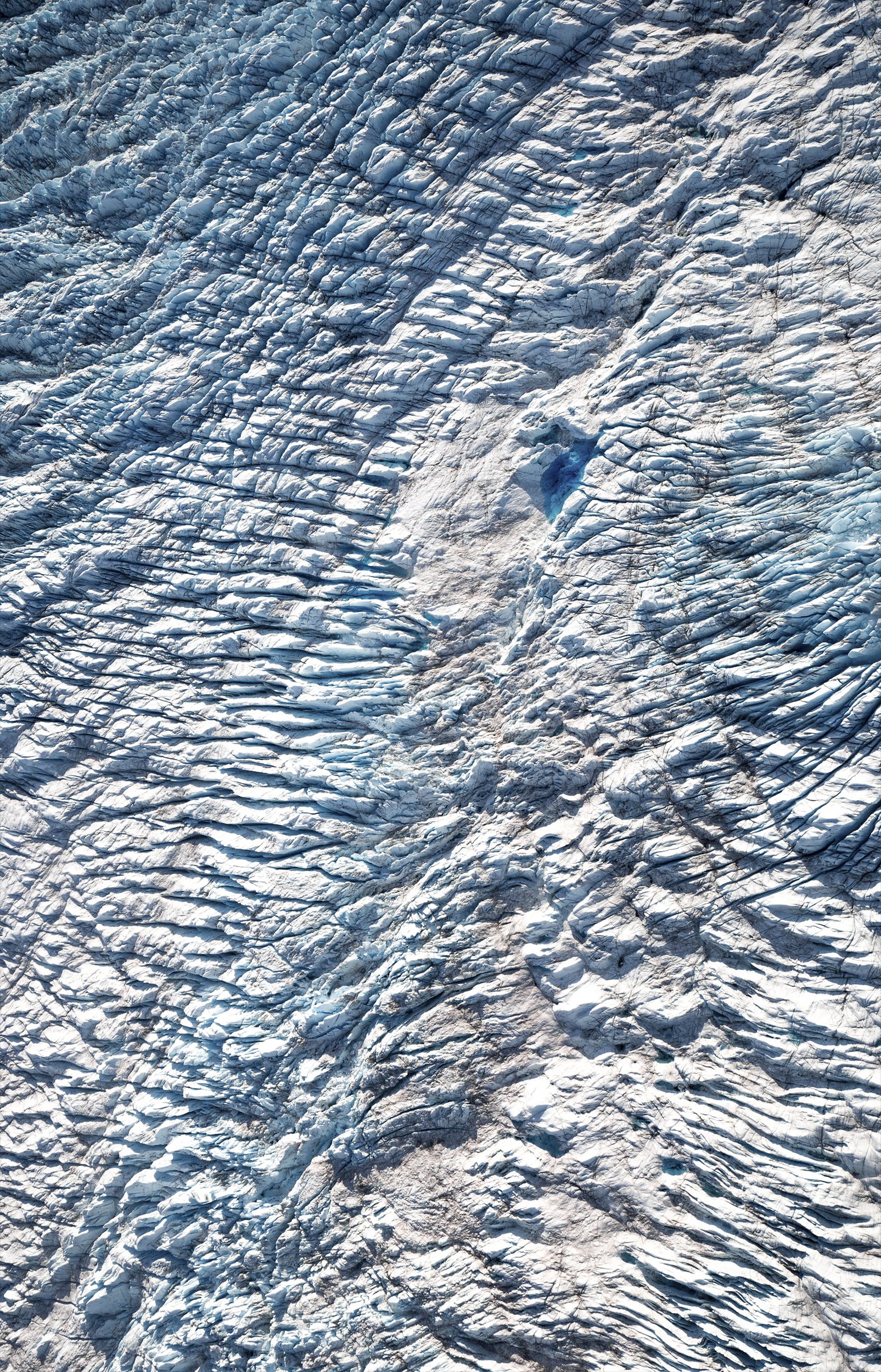 aerial view of glacier looking down at patterns in blue coloured ice. There is a small pool of turquoise blue water in the middle of the glacier.