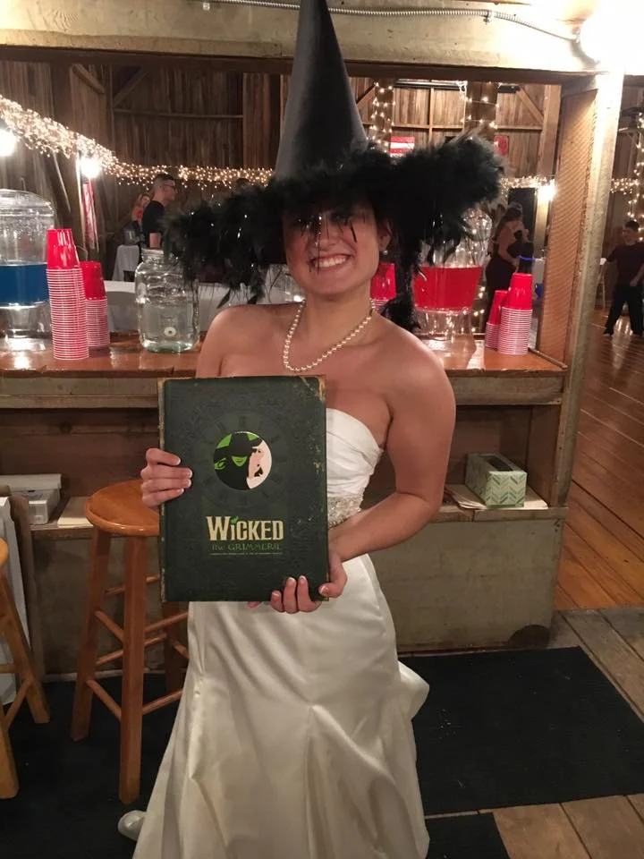 A bride wearing a wicked witch hat and holding a book from the Musical Wicked