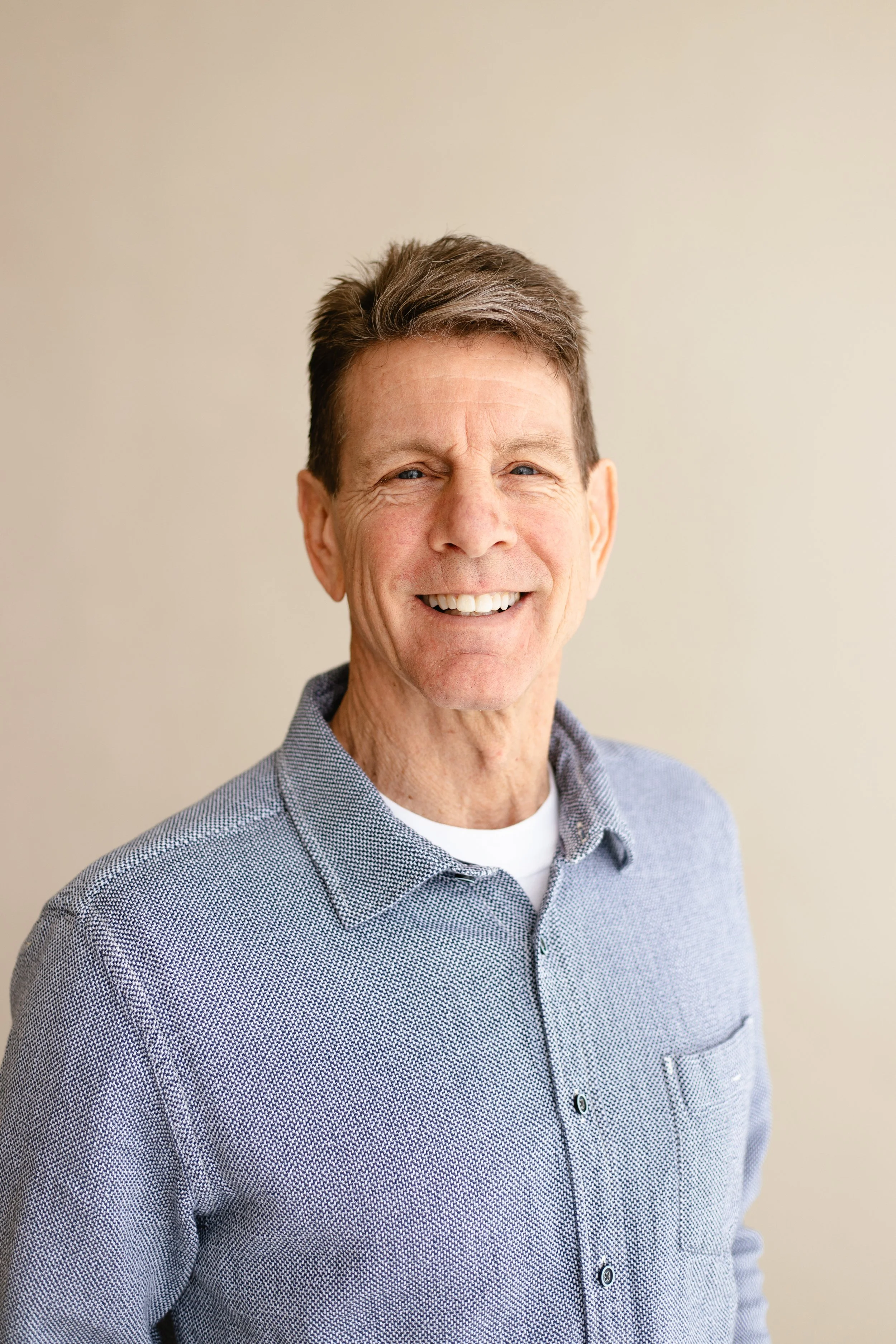 Portrait of a smiling middle-aged man with short brown hair, wearing a blue patterned button-up shirt against a plain beige background.