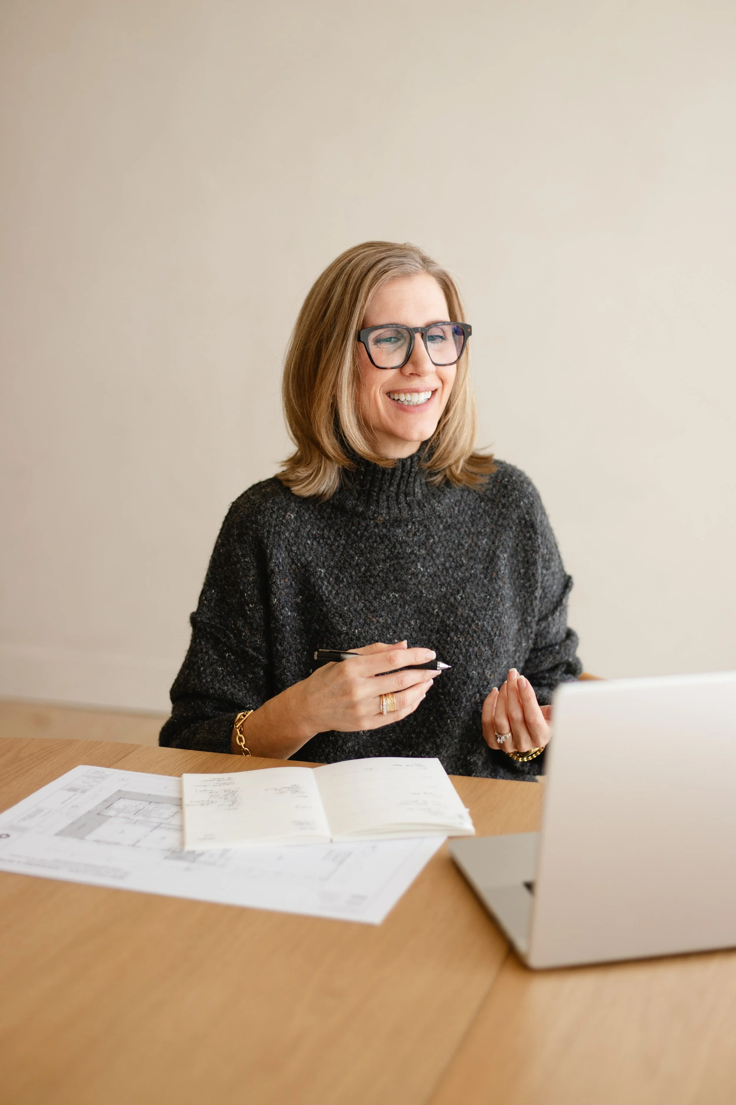 A woman with shoulder-length blonde hair, glasses, and a dark sweater sitting at a wooden table with papers and a laptop, smiling during a video call or meeting.