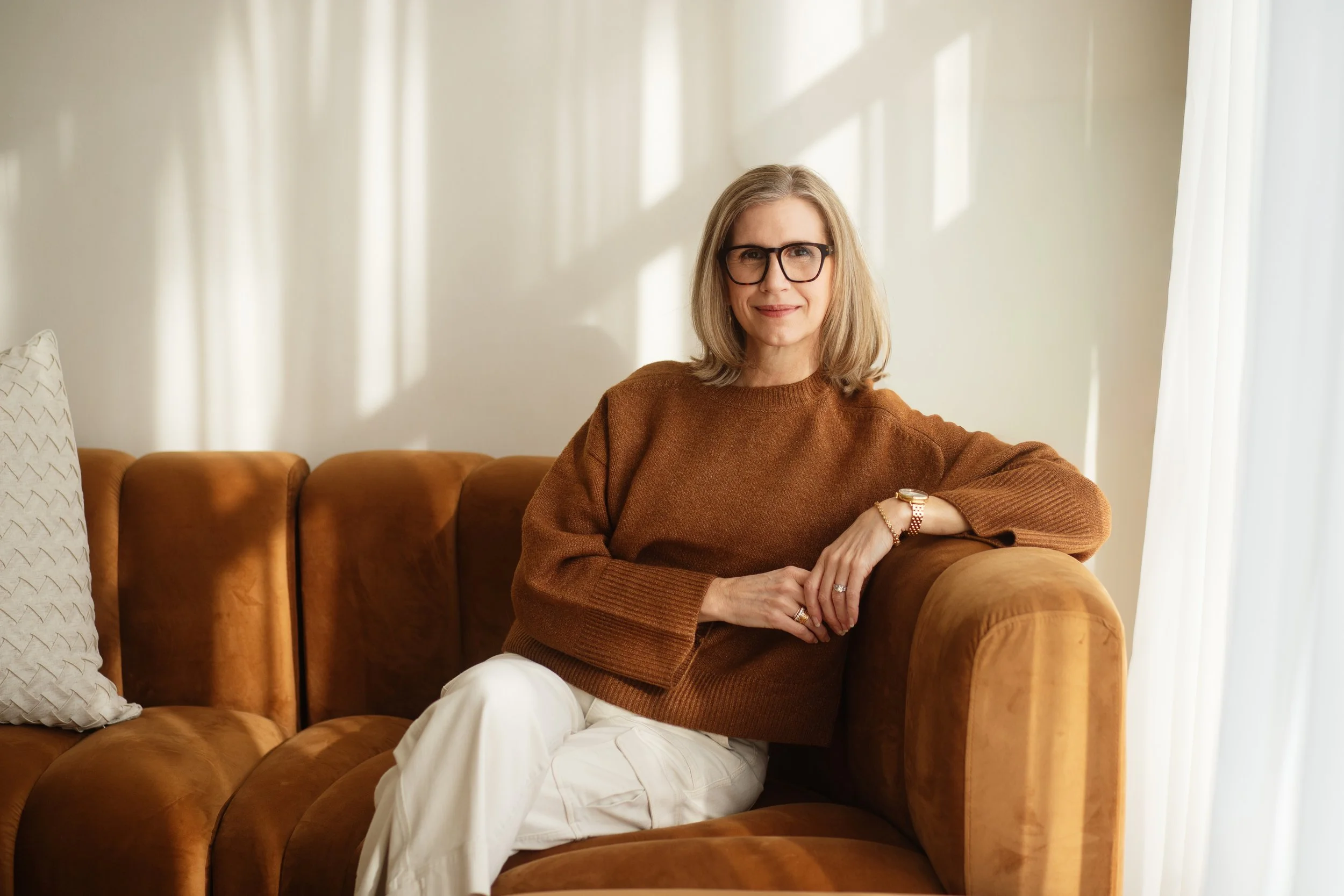 A woman with glasses and shoulder-length blonde hair sitting on a brown leather couch in a well-lit room, wearing a brown sweater and white pants.