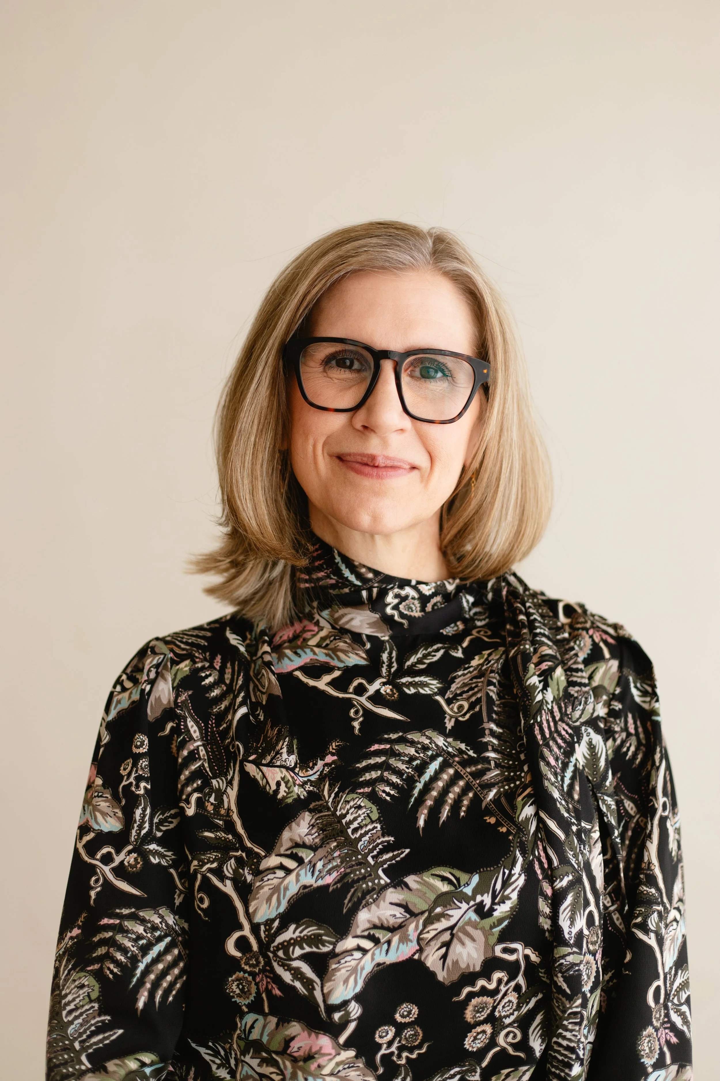 A woman with shoulder-length blonde hair and black glasses, wearing a black patterned blouse with a bow tie, standing against a plain light background.