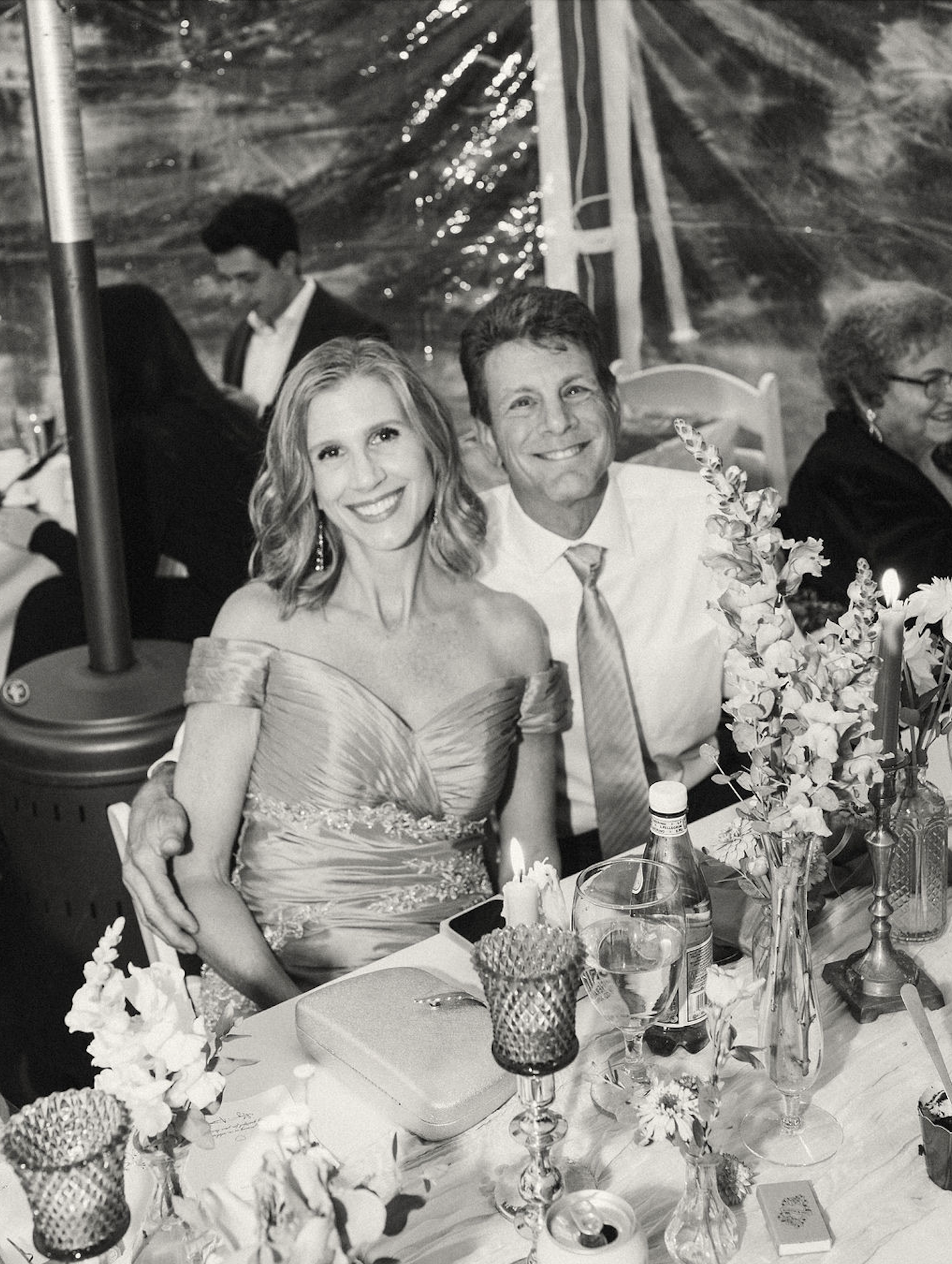 A smiling woman in an off-shoulder dress and a smiling man in a shirt and tie sitting at a decorated table with flowers, candles, and drinks, at a celebration event.