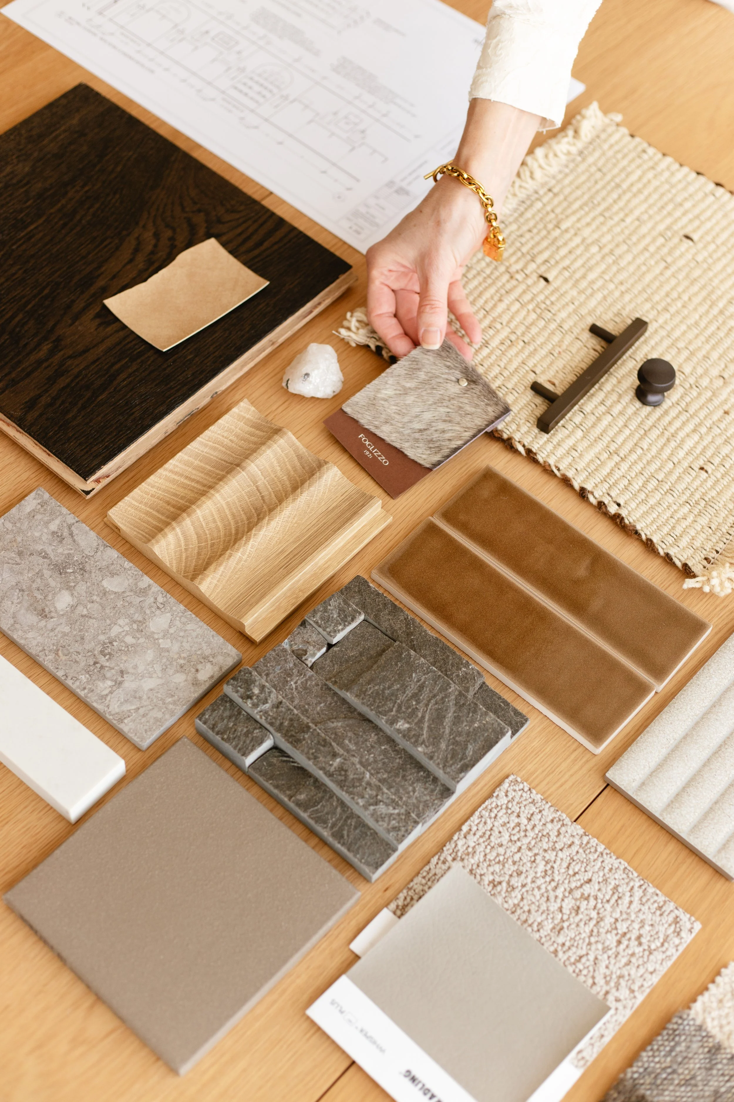 A person arranging various material and color samples, including tiles, fabric swatches, and wood textures, on a wooden table.