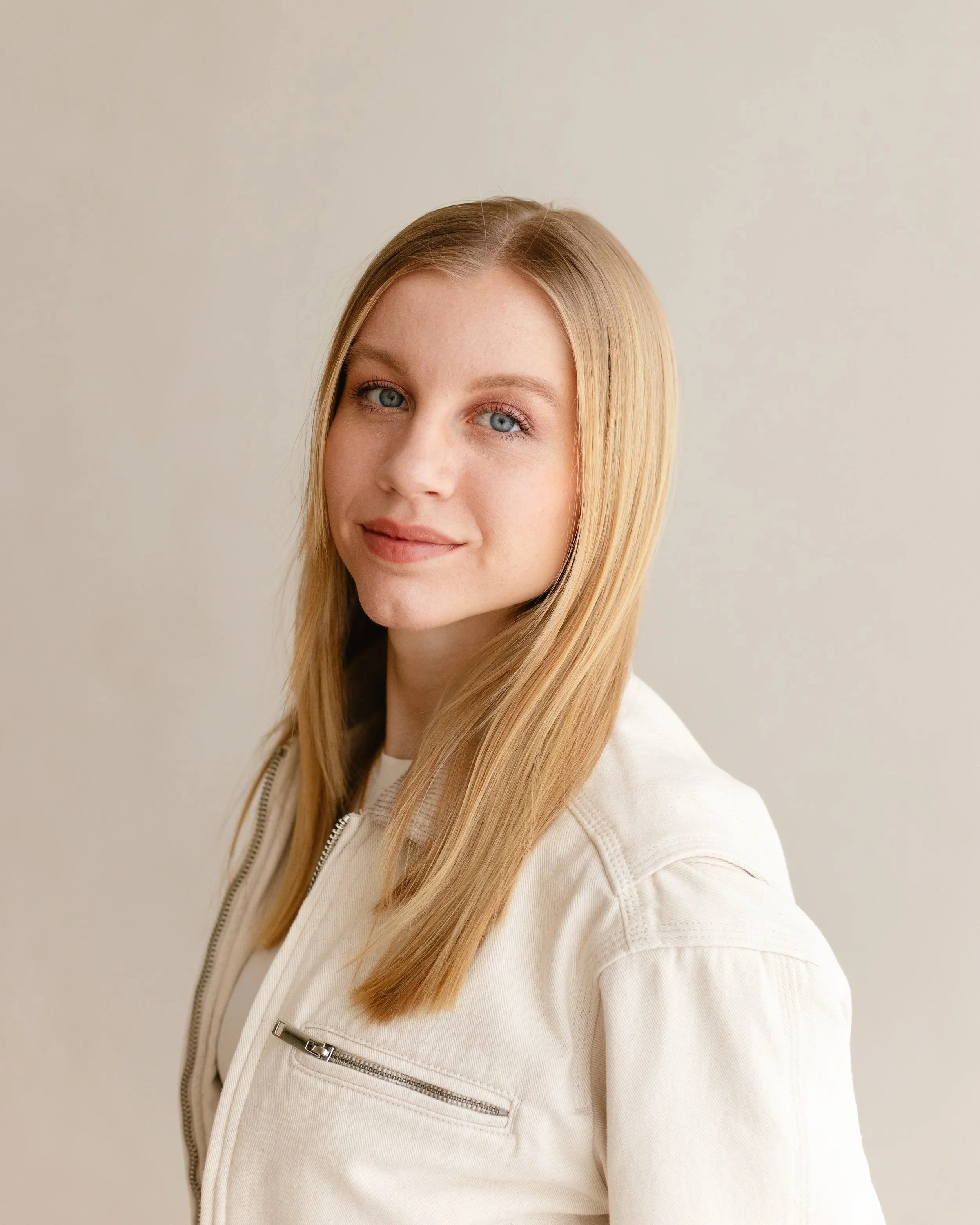 A young woman with long blonde hair, blue eyes, and fair skin, wearing a white jacket, standing against a plain light-colored background.