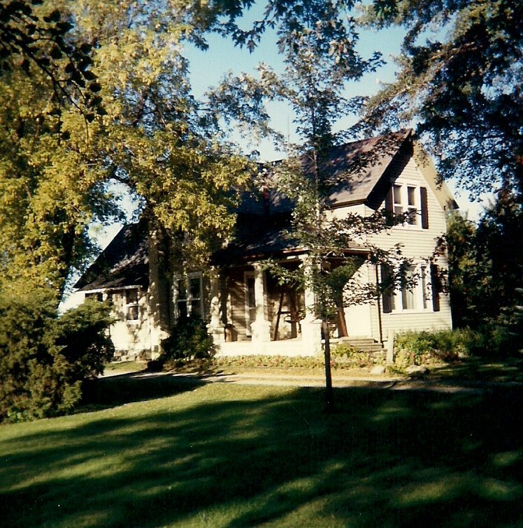 A two-story house with a front porch, surrounded by trees and a grassy lawn, under a clear blue sky.