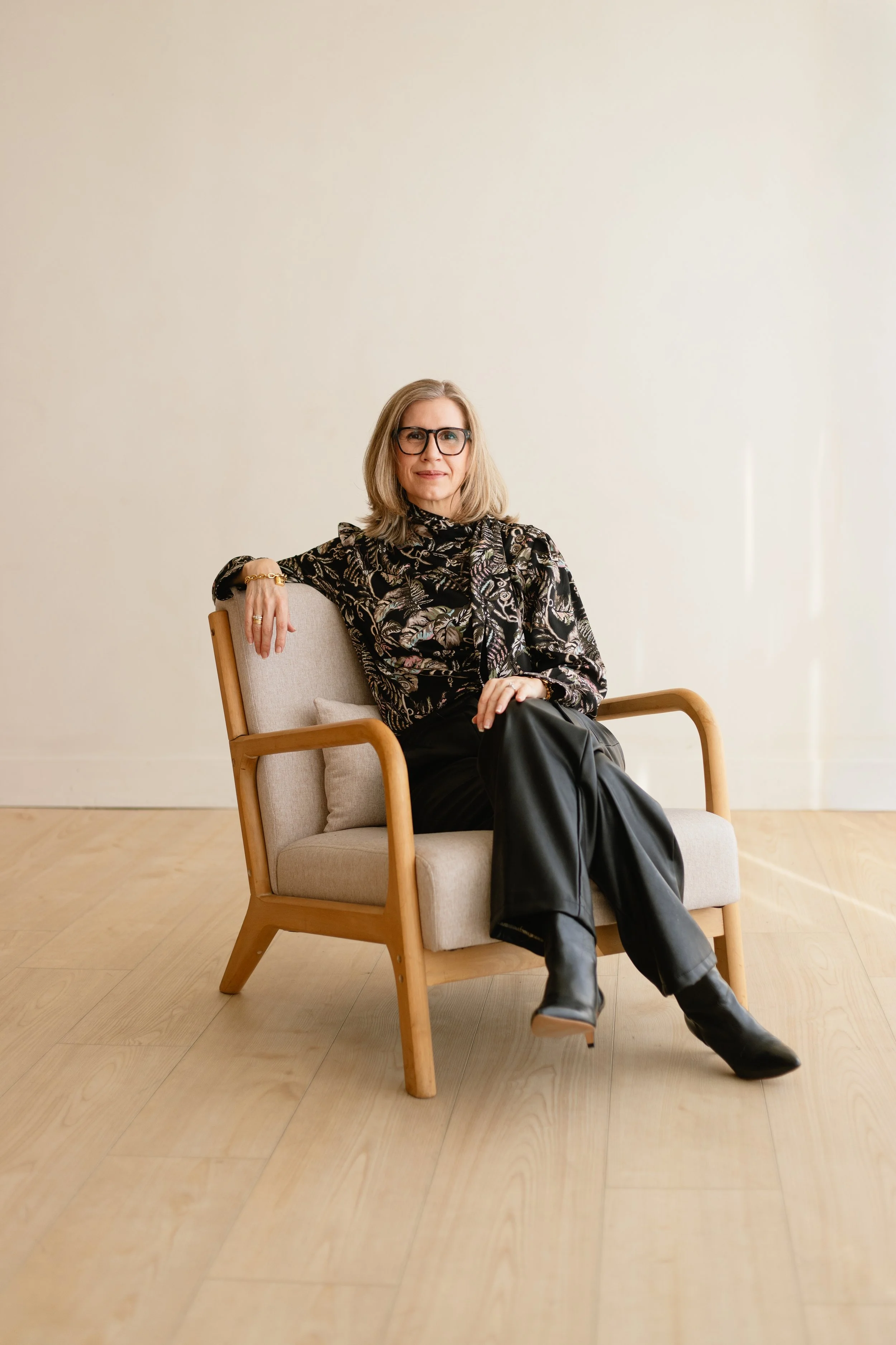 A woman with shoulder-length blonde hair and glasses sitting on a beige armchair with wooden arms, wearing a patterned black and gold blouse, black leather pants, and black boots, in a room with light wood flooring and a plain light-colored wall.