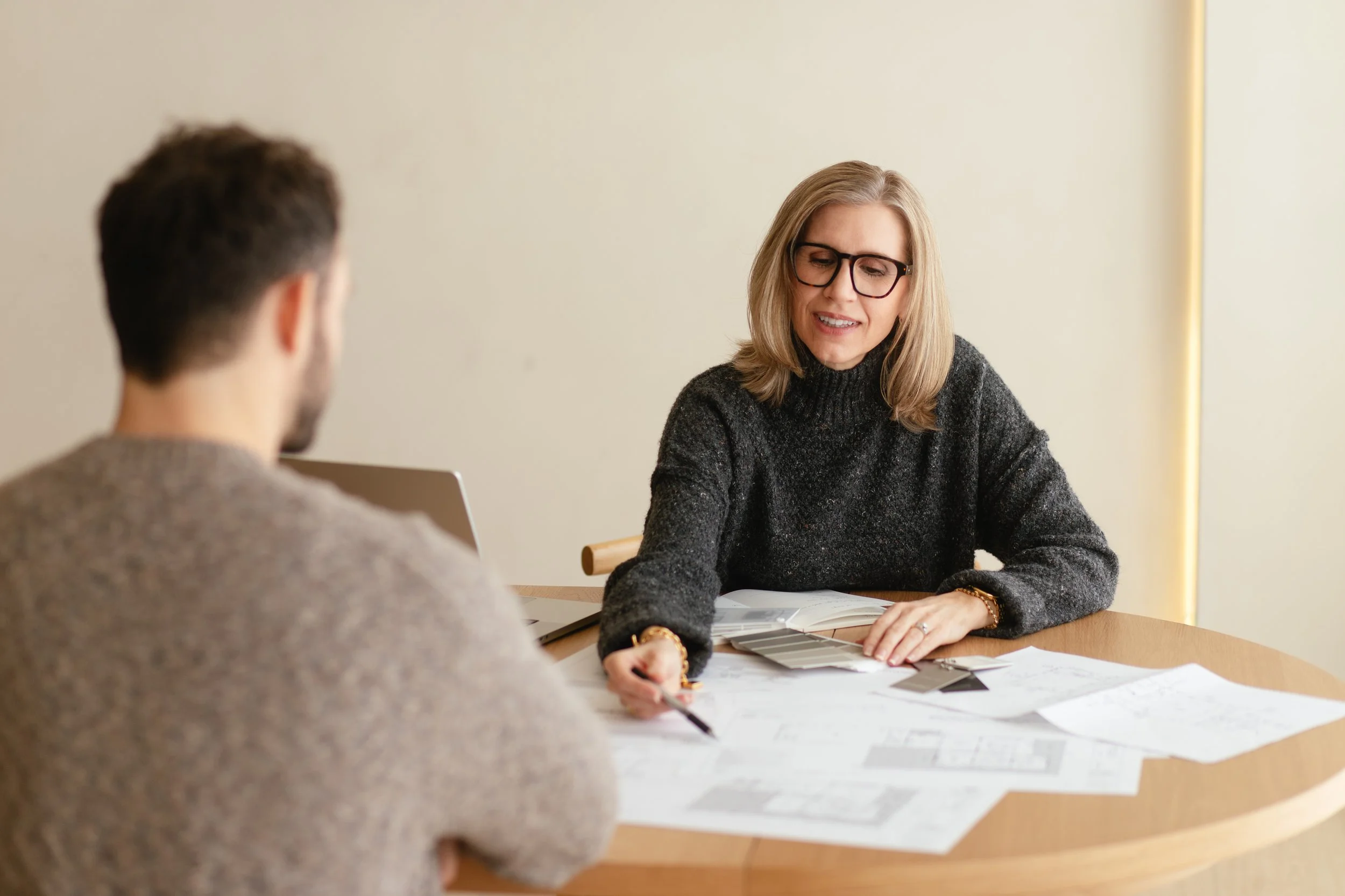 A woman with glasses and blonde hair wearing a dark sweater sitting at a table with a man, discussing documents and color swatches in an office setting.