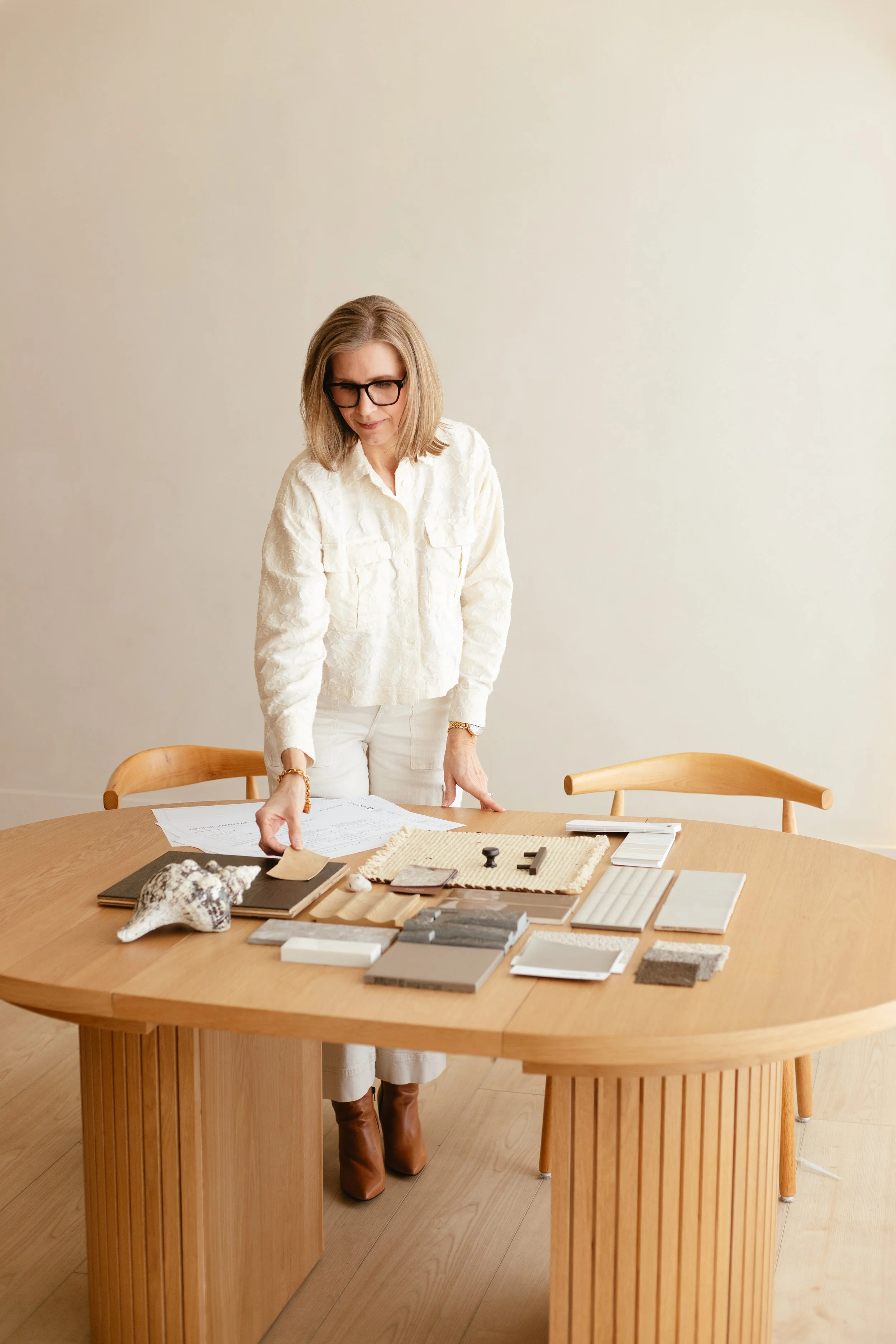 A woman in white clothing and brown boots is standing at a wooden table with design samples, fabric swatches, and documents, in a minimalist room with a plain wall.