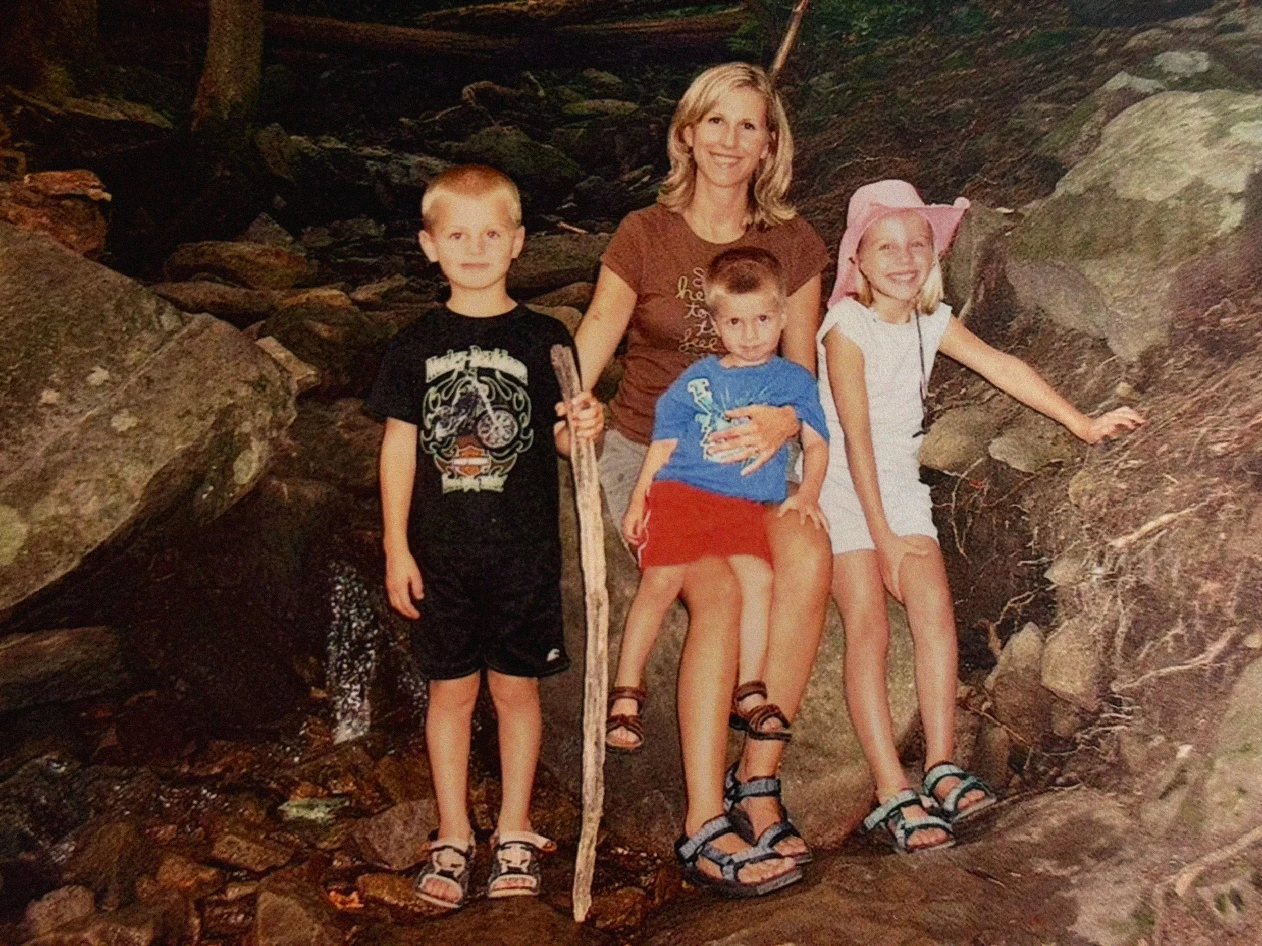A woman with three children in a rocky outdoor setting, one girl with a pink hat, two boys, one holding a stick.