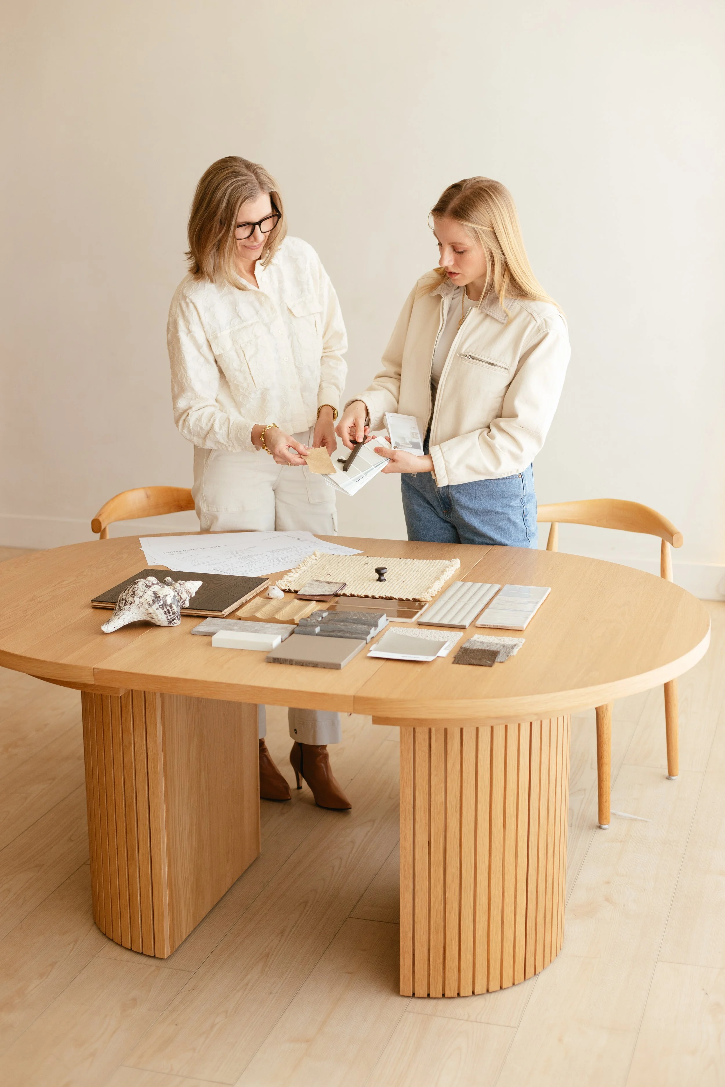 Two women discussing fabric samples, paint swatches, and design materials spread out on a wooden table in a bright room.