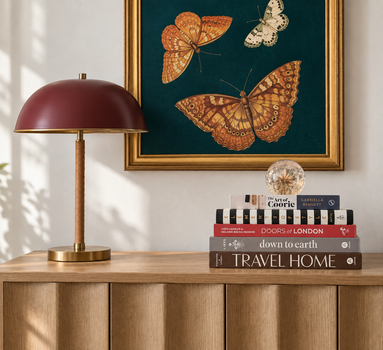 Clear botanical paperweight with a preserved plant inside, styled on stacked books beside a burgundy table lamp on an oak console by virtual interior designer Joshua Jones of JJones Design Co.
