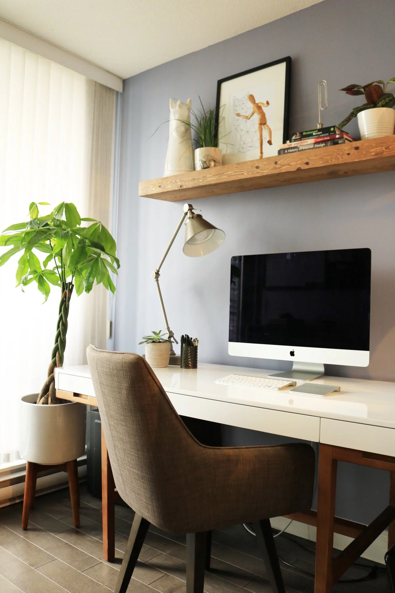 A modern home office designed by virtual interior designer Joshua Jones in his former Vancouver, BC apartment, featuring a white desk, iMac computer, wood shelving, indoor plants, and warm natural lighting.