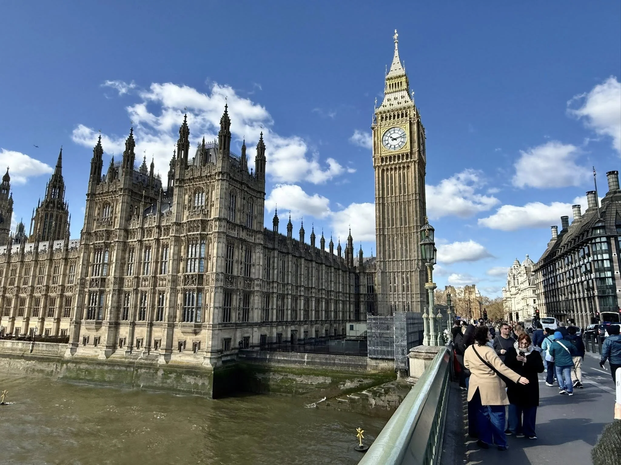 Big Ben and the Houses of Parliament in London viewed from Westminster Bridge with the River Thames and pedestrians in the foreground under a clear blue sky