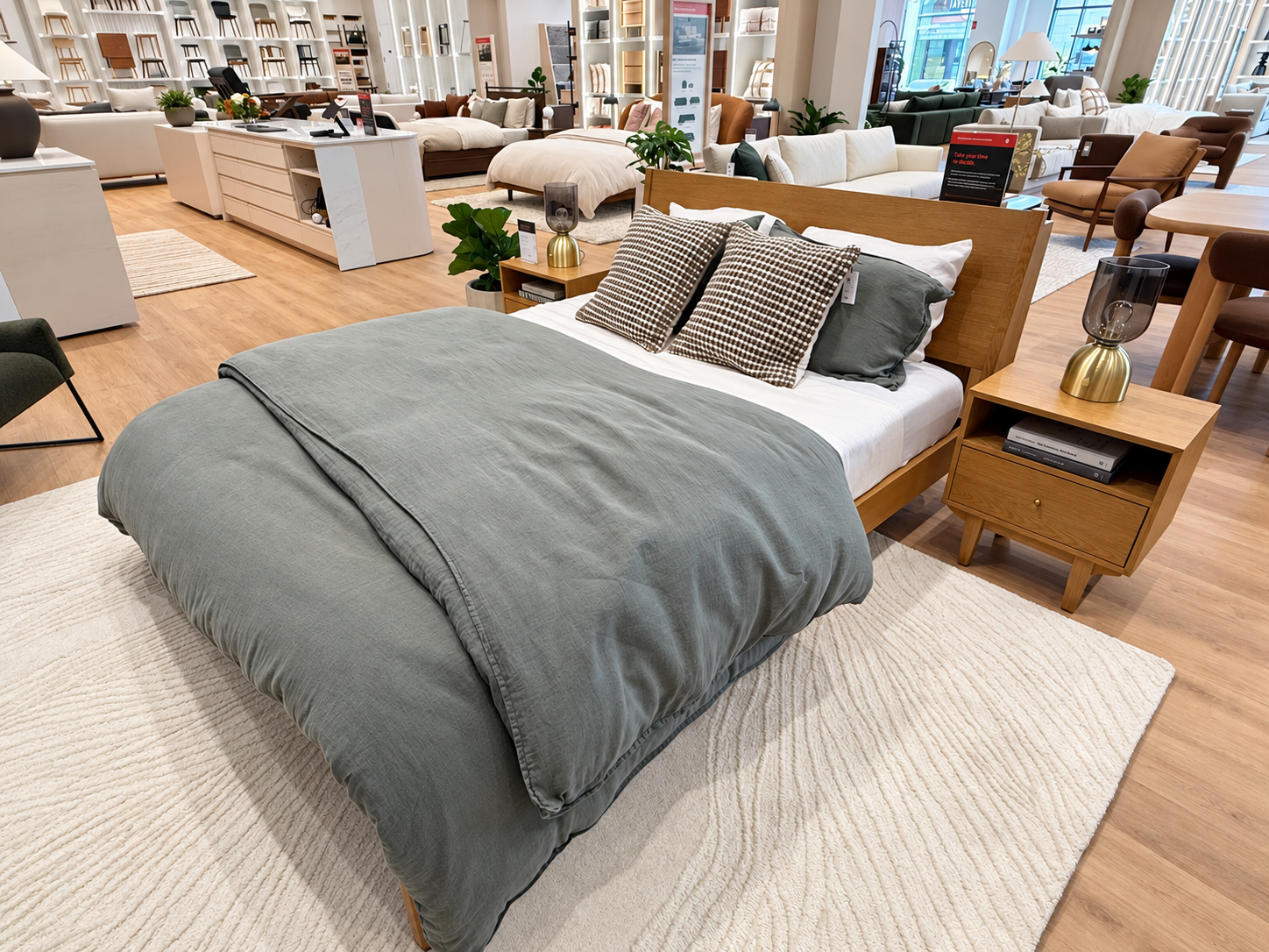 Wood bed with sage-gray bedding and checked pillows, flanked by matching nightstands with brass and smoked-glass lamps on a textured light-colored rug.